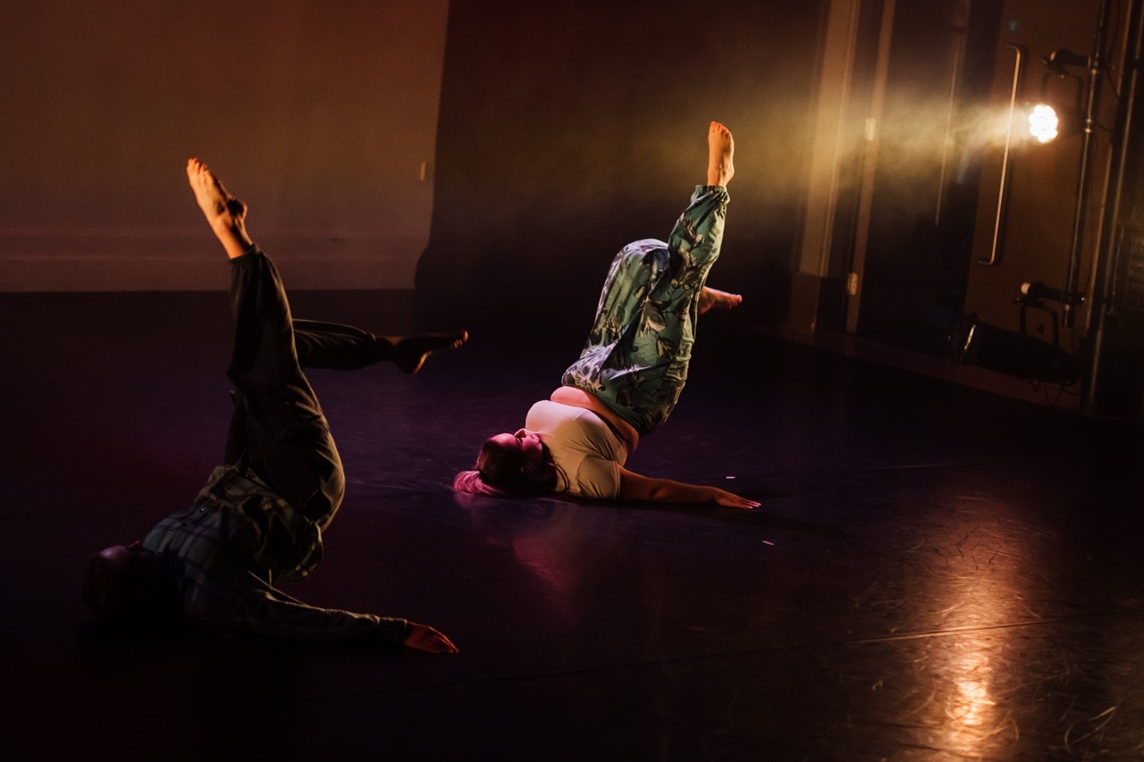 Two dancers performing on dark wooden floor with dramatic stage lighting creating warm orange glow and shadows in studio space.