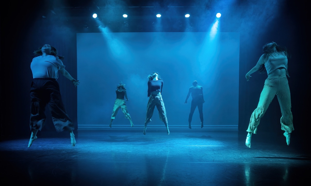 Five dancers on stage under bright blue spotlights, with silhouettes visible in background. Dark theatre setting with dramatic lighting.