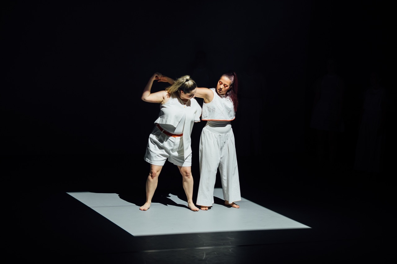 Two performers in white clothing on illuminated square platform against black background, one kneeling, one standing.
