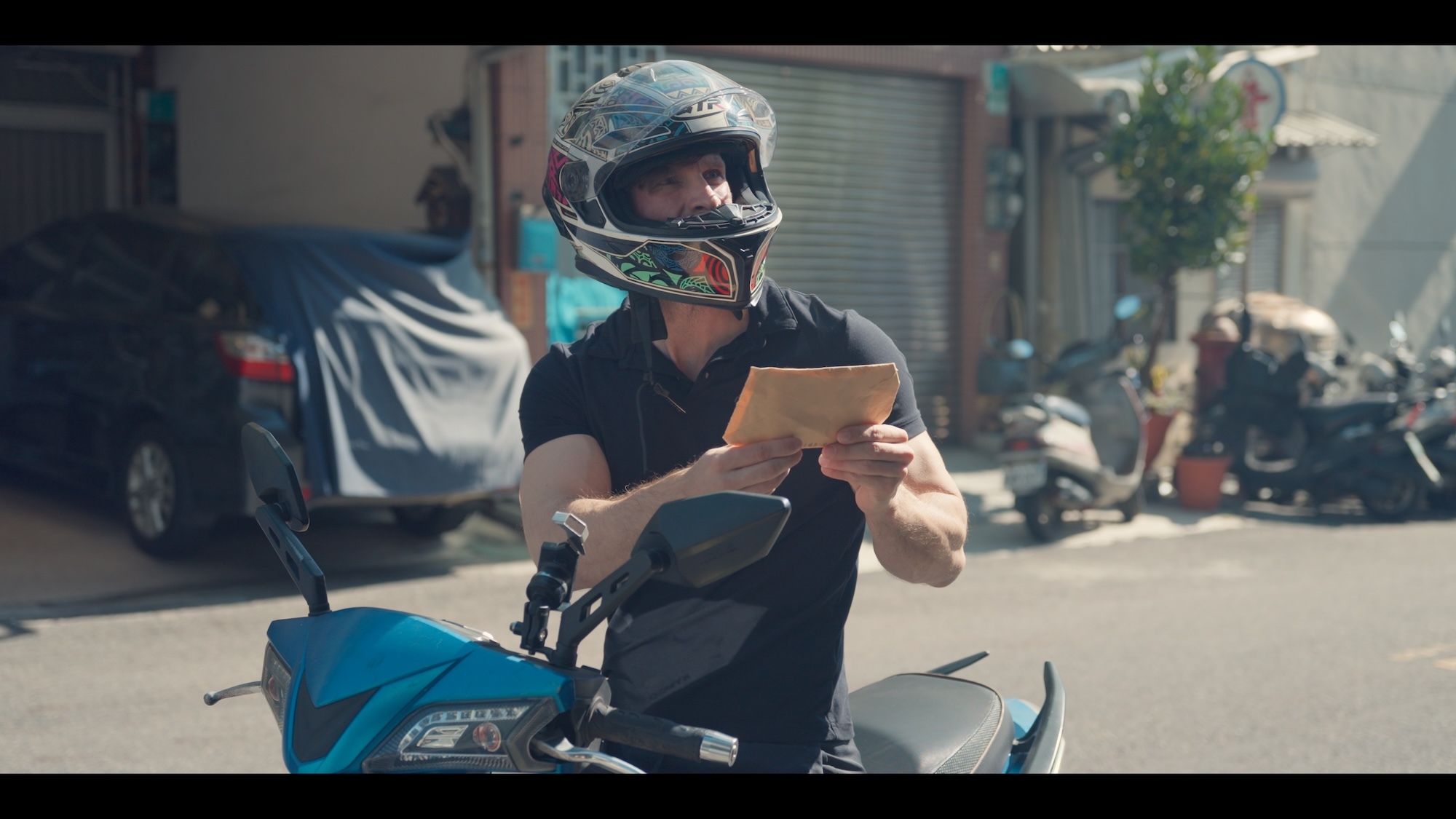 Still from the short film "Found and Forgiven", showing actor and filmmaker P Kertesz on a motorbike wearing a helmet in a Taiwanese street, holding a piece of paper and looking up at a building off screen.