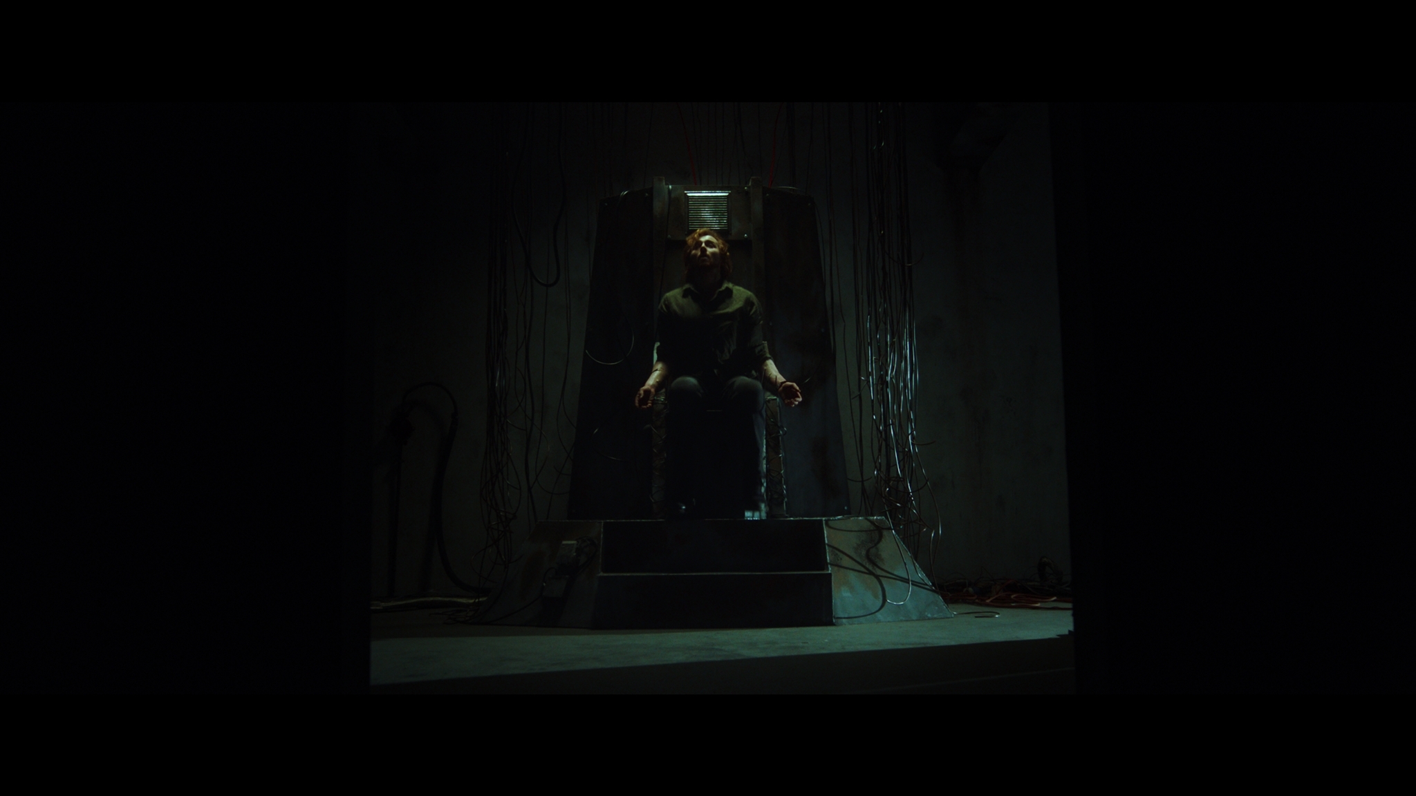 Dark interior with figure in black clothing seated in centre, illuminated by green light against shadowy background.