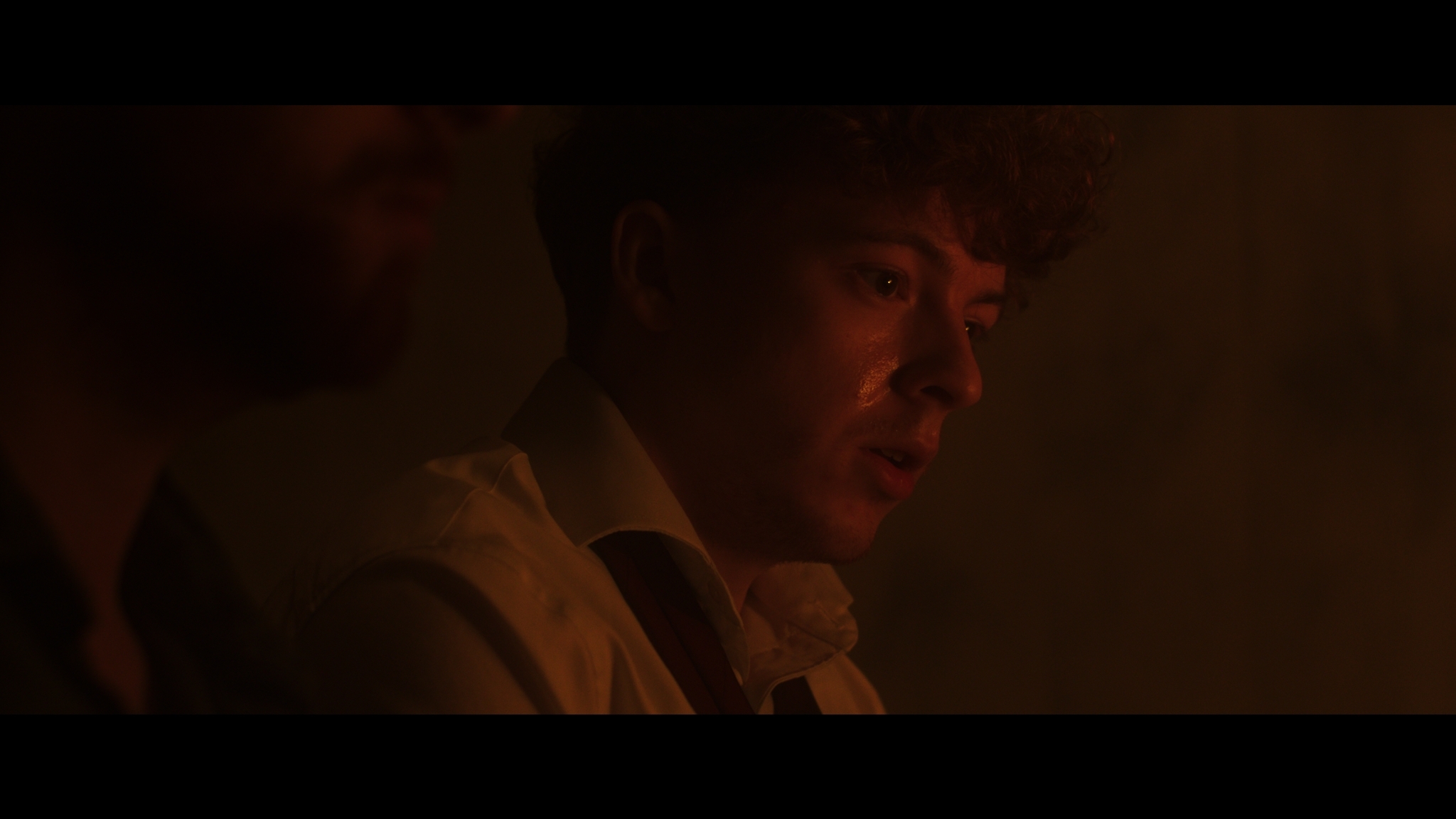Man in dark room lit by warm orange light, face partially visible in shadow, wearing light-coloured jacket.