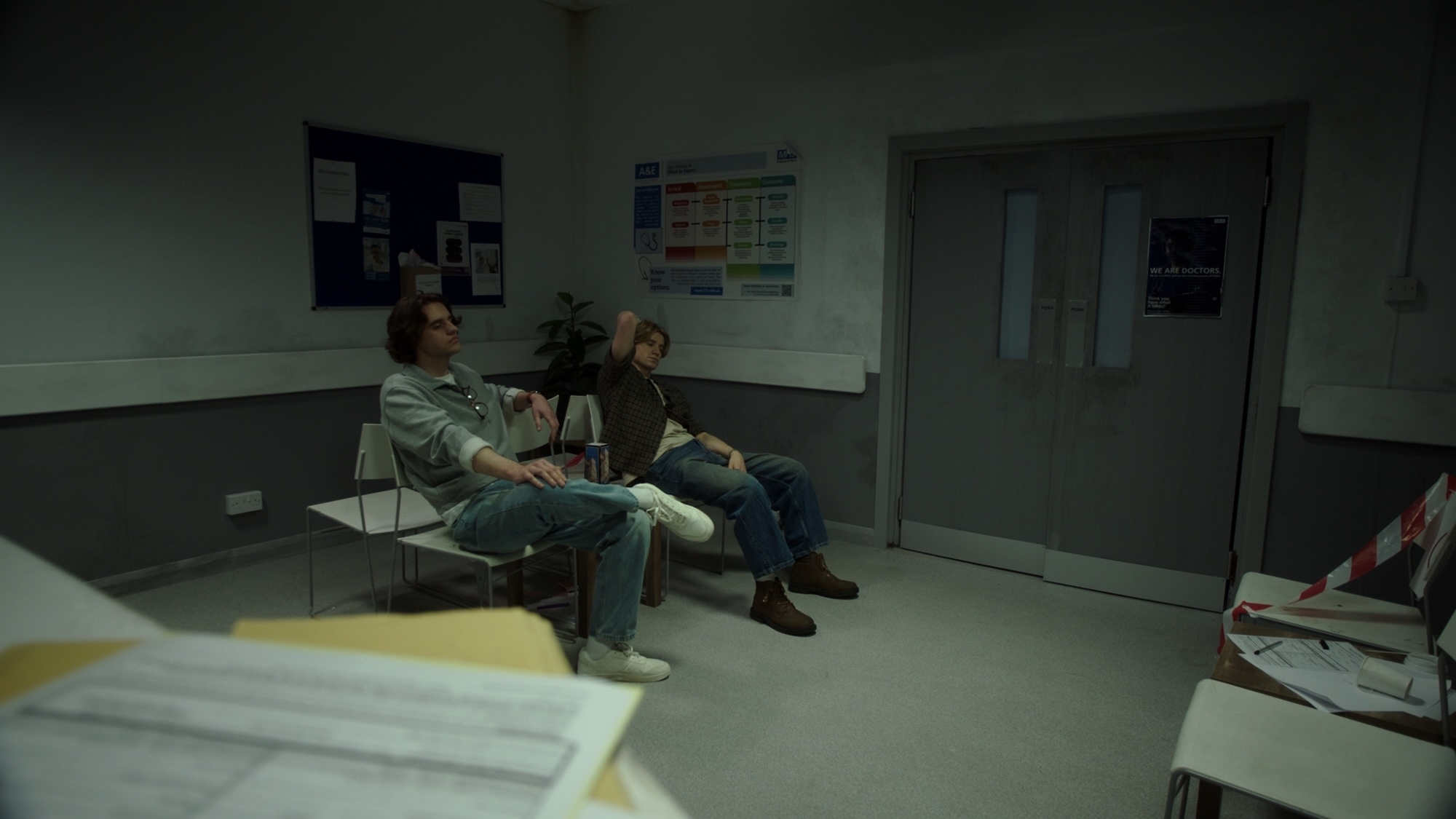 Two people sitting in hospital waiting area with grey and green walls, metal chairs, and fluorescent lighting.