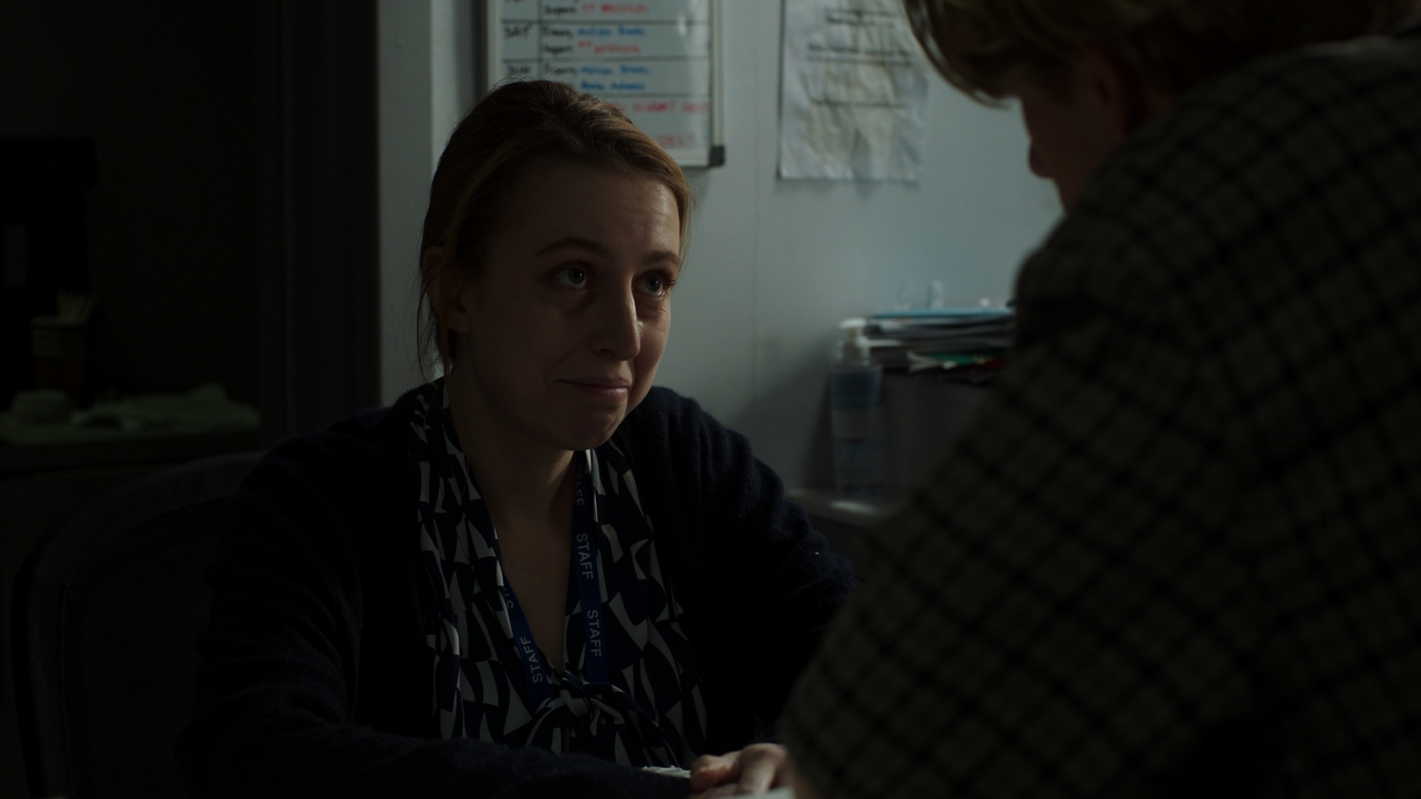 Close-up of a person in patterned dark shirt sitting at desk facing person in office setting with papers on wall behind them.