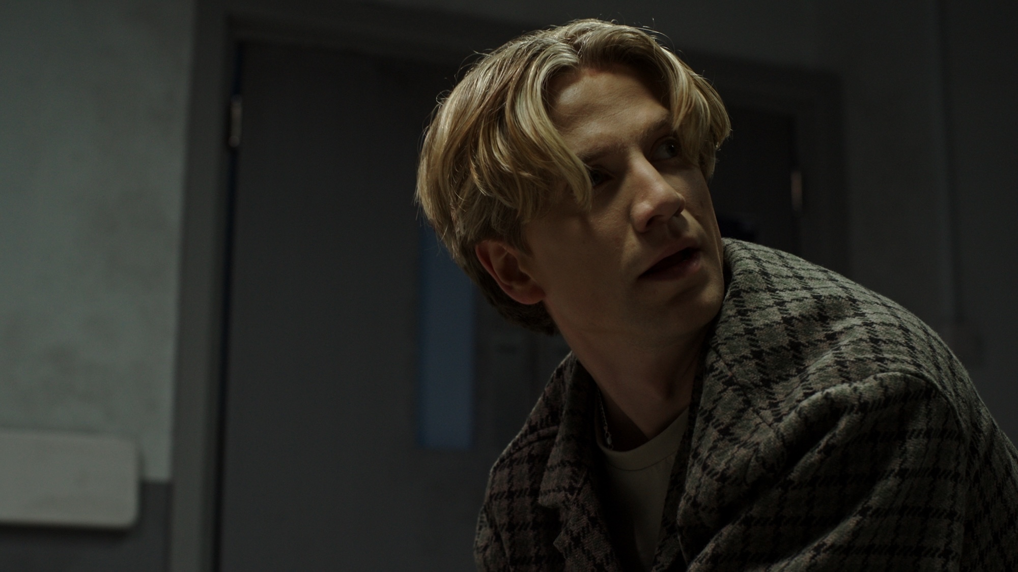 Person with blonde hair wearing grey checkered shirt in dimly lit room with dark background.