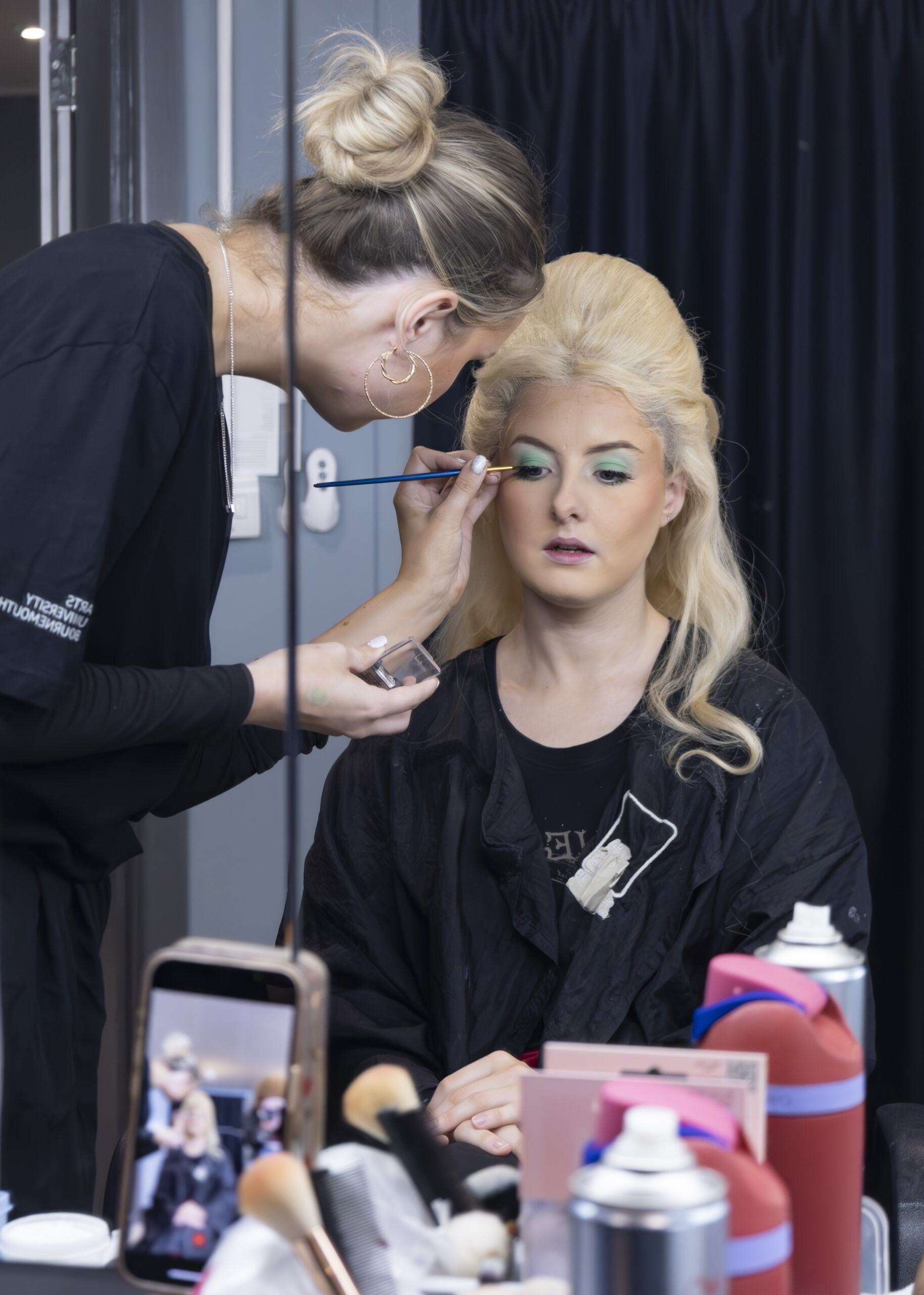 Makeup artist with blonde hair in bun applies makeup to blonde woman in black top, with cosmetics and mobile phone on counter.