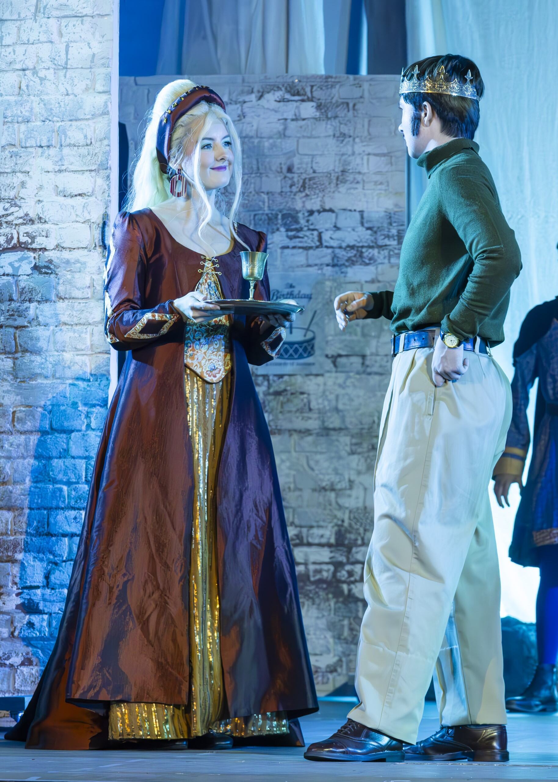 Woman in burgundy medieval gown with white headdress faces man in green military jacket and white trousers on stage with brick backdrop. Blue stage lighting.