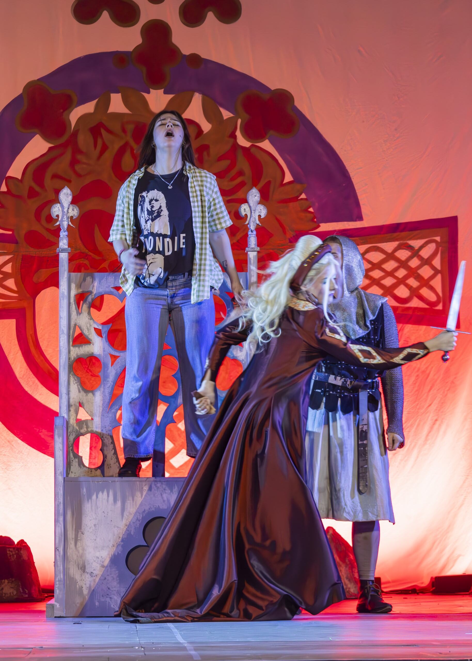 Person in casual clothes standing on platform with ornate red backdrop, whilst costumed figure in dark robes kneels below on stage.