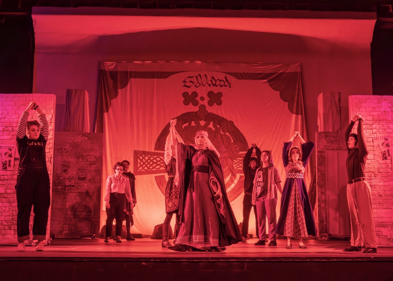 Theatre stage bathed in red lighting with performers in period costumes. Central archway with ornate set design and columns.