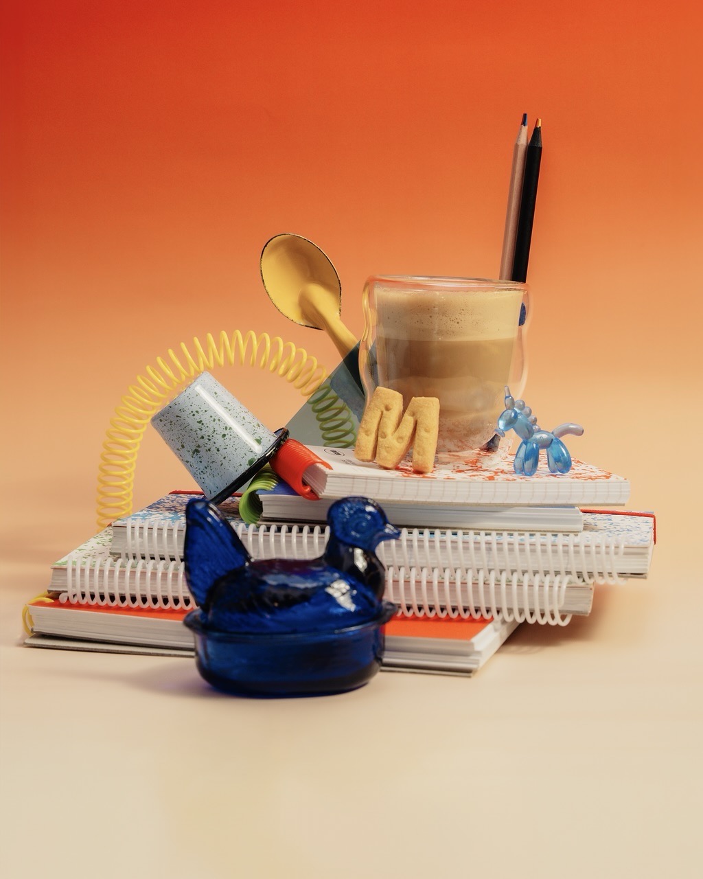 A stack of notebooks with coiled spiral bindings, topped with a glass cup, pencils, a spoon and scattered small decorative objects on a cream surface against an orange background.