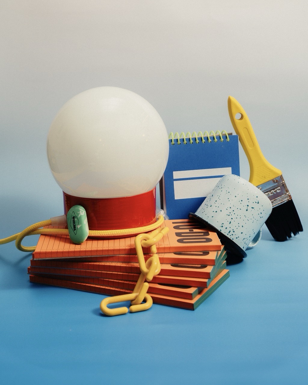 A desk organiser with a white globe lamp, stacked notebooks and stationery items, including a yellow paintbrush and a blue taped roll, on a blue surface.