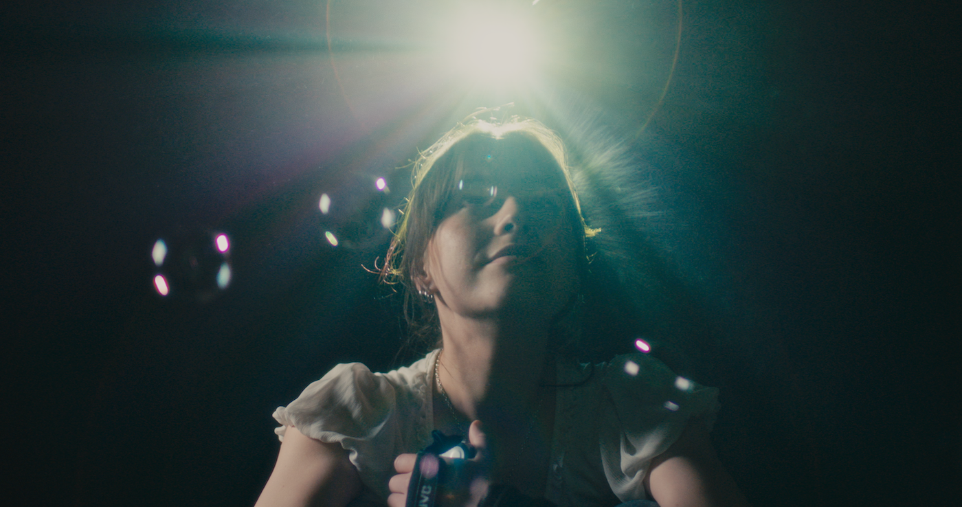 Film still from "Time Between Us" – person with dark hair and white top in dim lighting with coloured lens flares and light spots scattered across dark background.