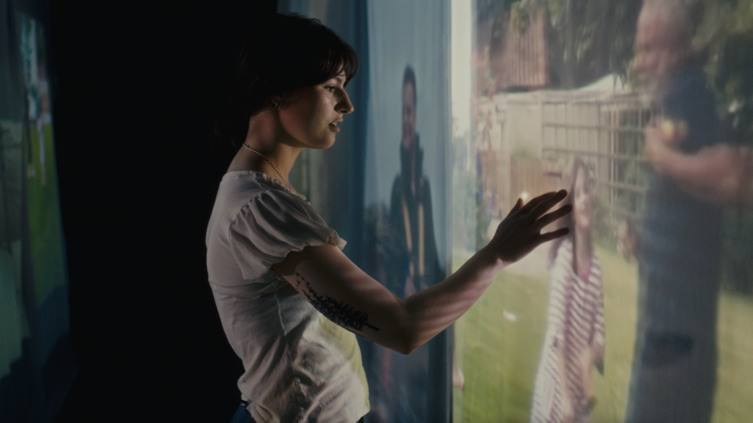 Film still from Time Between Us (2024) – person in grey T-shirt touching translucent digital display with text and graphics, colourful projected light on walls.