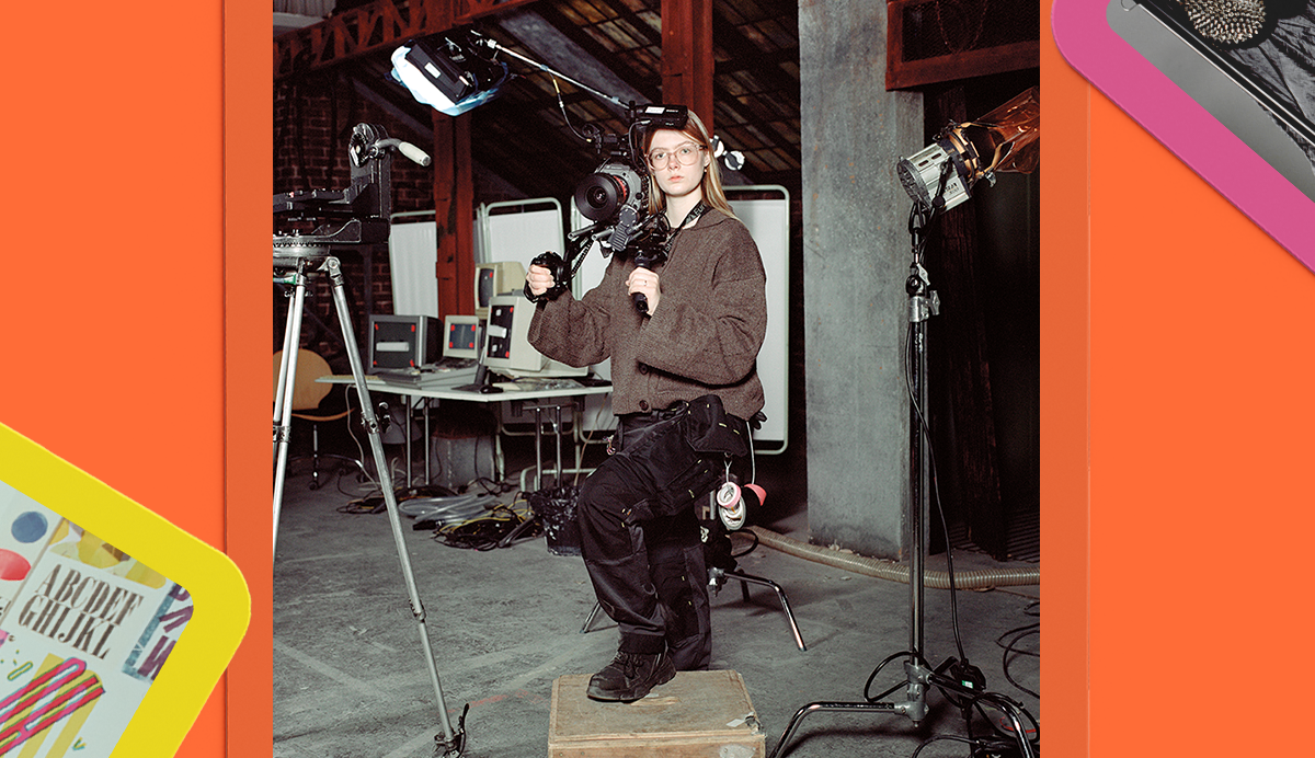 Man in brown jumper sits in television studio with cameras, tripods and equipment. Orange and pink geometric shapes frame image.