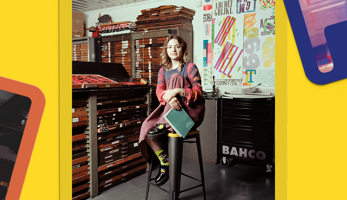 Person with long hair sits on stool in colourful classroom with wooden storage shelves, educational posters, and yellow walls.