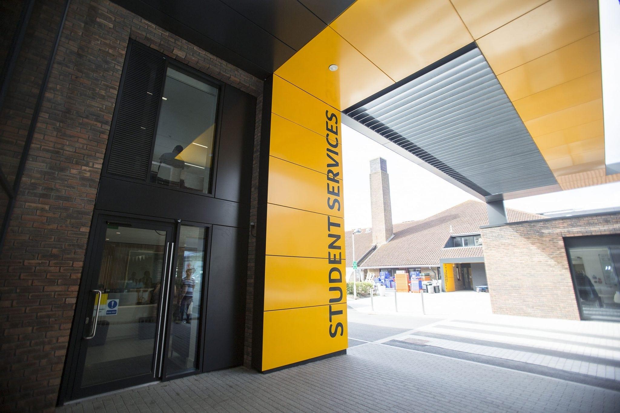A university building with a large doors and windows. There are yellow panels on the wall with Student Services written vertically across them.
