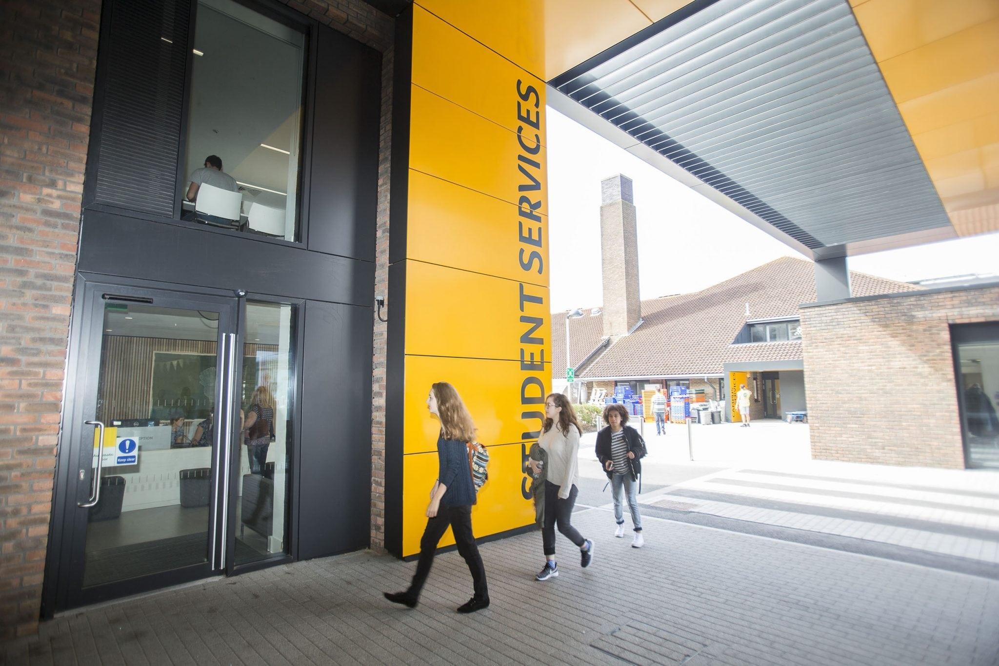 Students walking into a university building. There are yellow panels on the wall with Student Services written vertically across them.