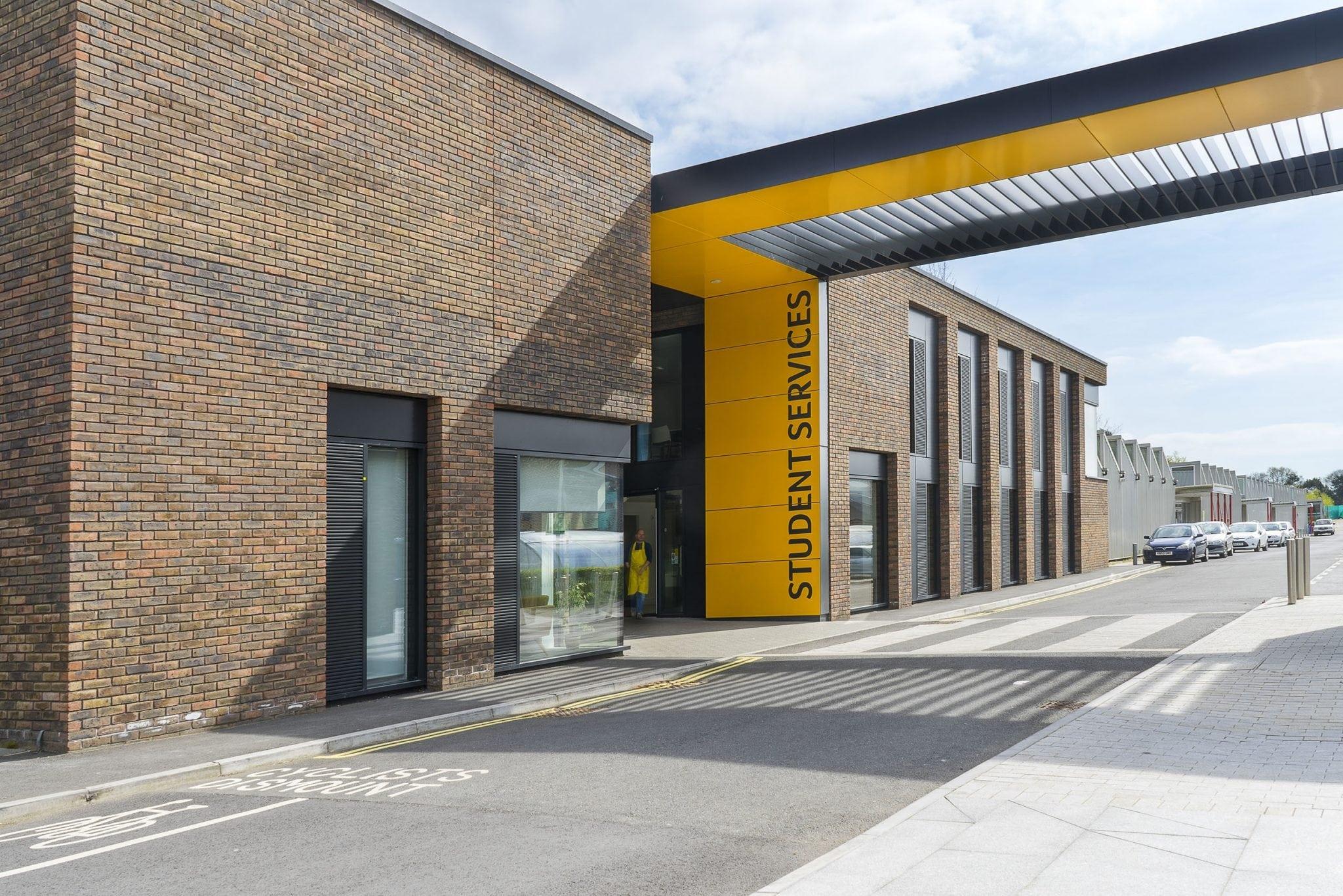 A brick university building with a road running alongside it. The road passes under a large rectangular archway with yellow panels on it. The words 'Student Services' appear on the archway.