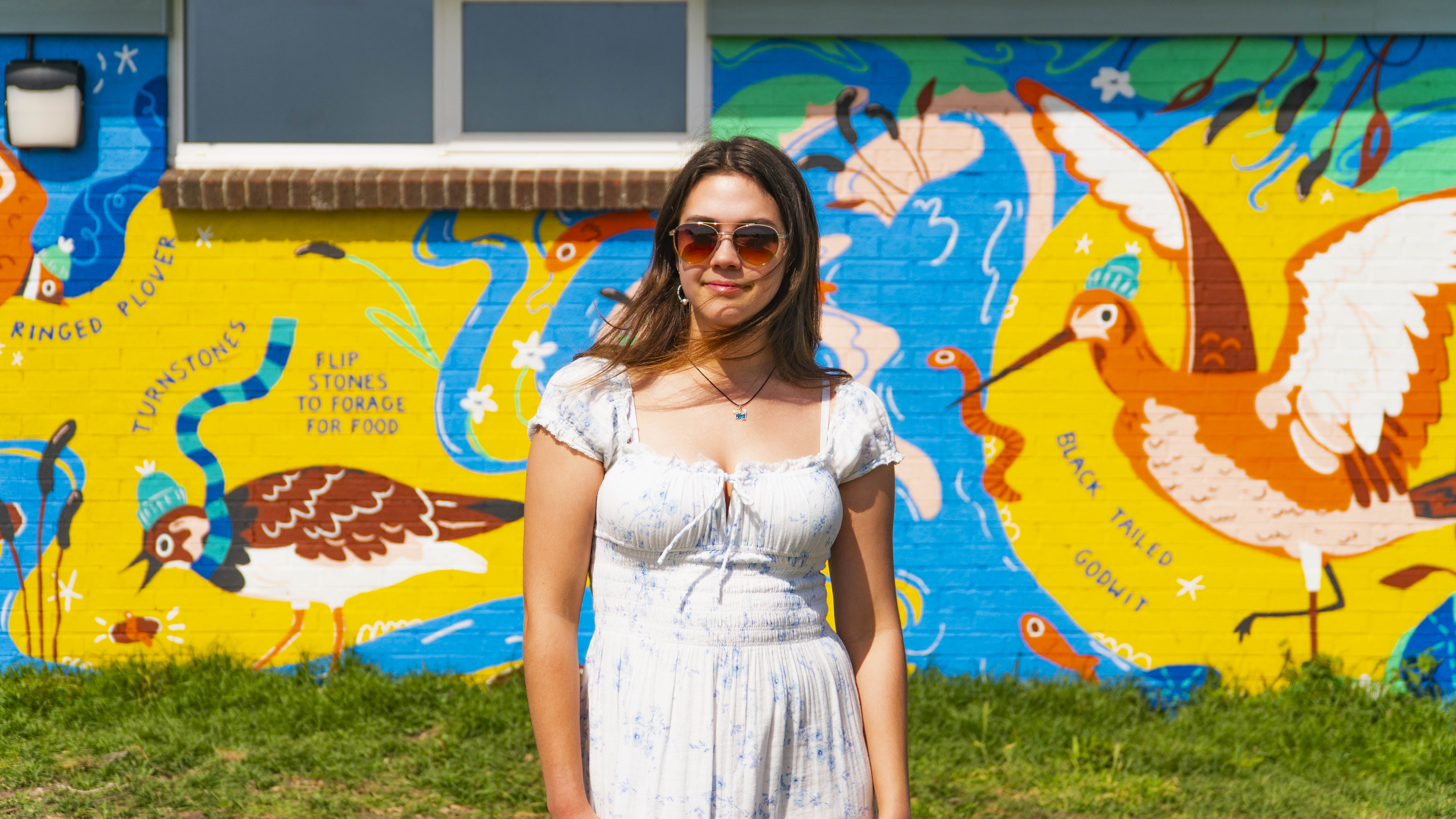 BA (Hons) Illustration student S Smith stands and poses for a photo in front of a bold and colourful mural depicting bird species on a building behind her.