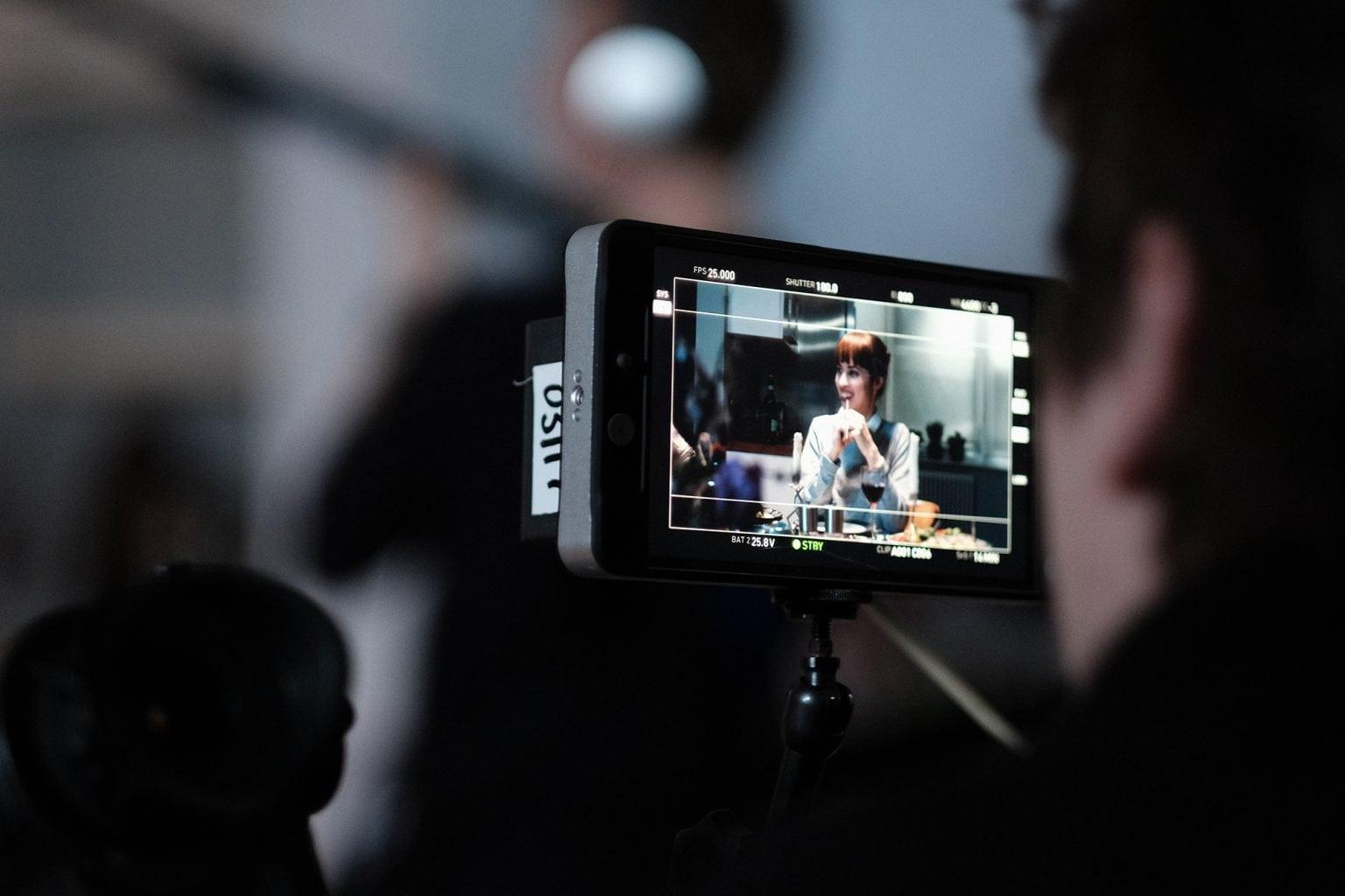 Close-up of a camera monitor while a scene is being filmed. A woman sat at a table with a glass of wine is being filmed.