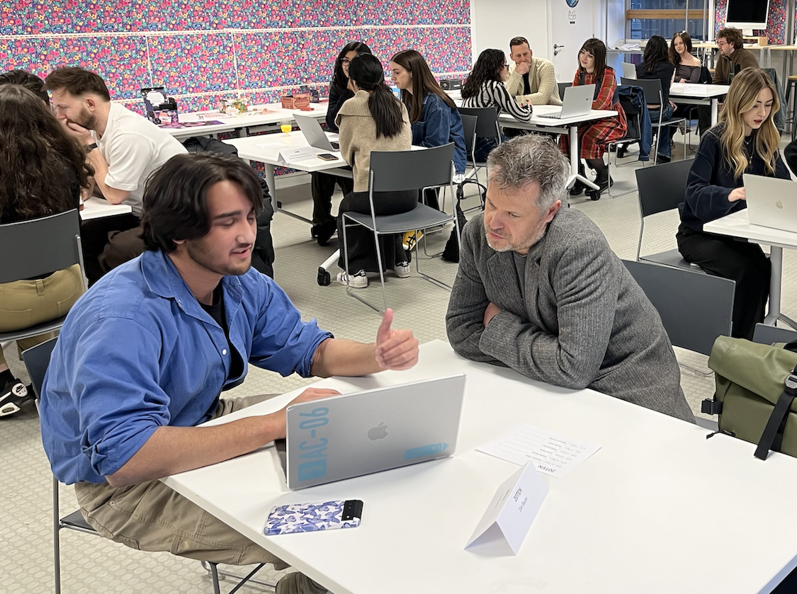 BA (Hons) Graphic Design students sitting at white tables in modern classroom with colourful patterned wall. A student sits with D Bacon in foreground discussing over laptop.