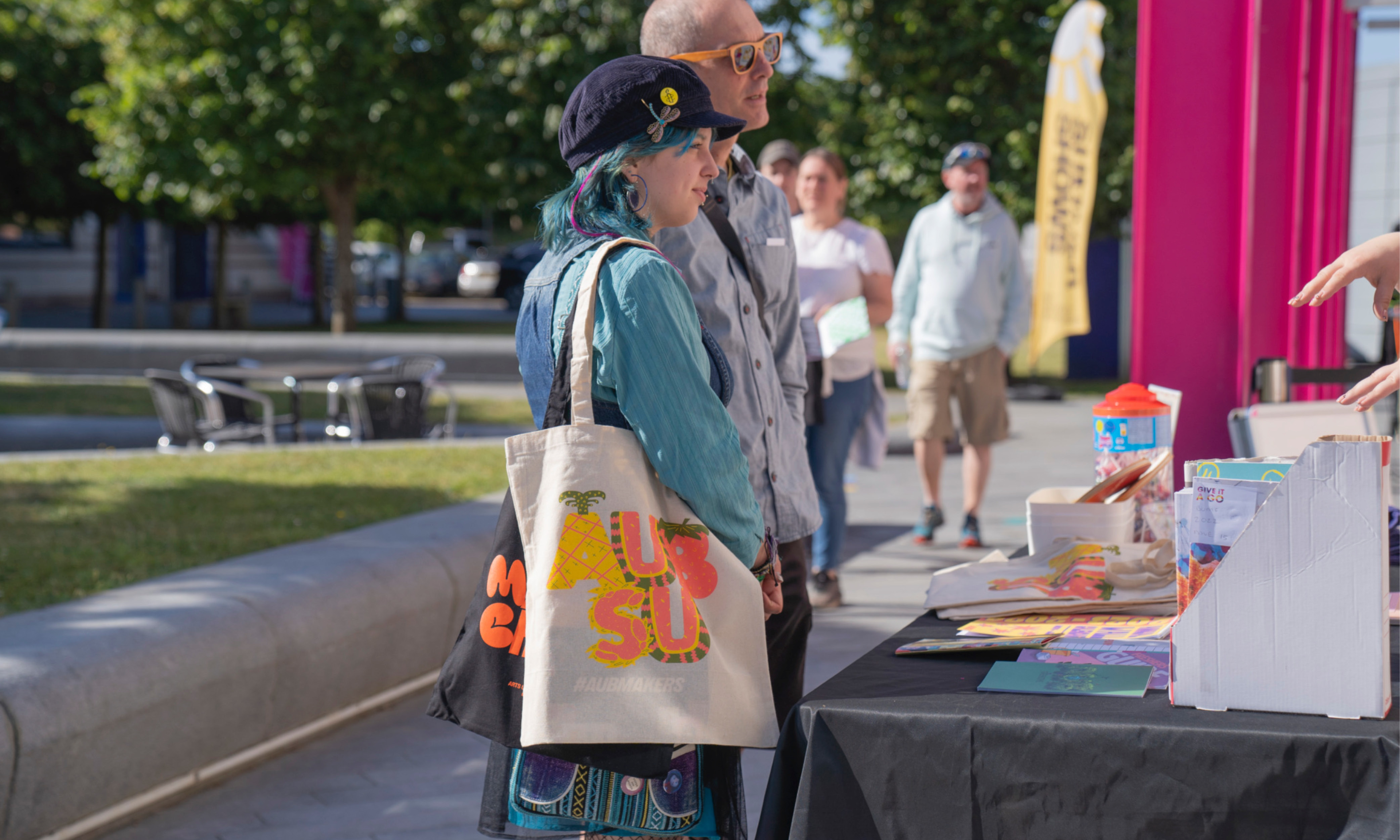 Student with parent, wearing blue clothing and a summer-themed orange AUBSU tote bag, stood outside in AUB Campus on a sunny morning.