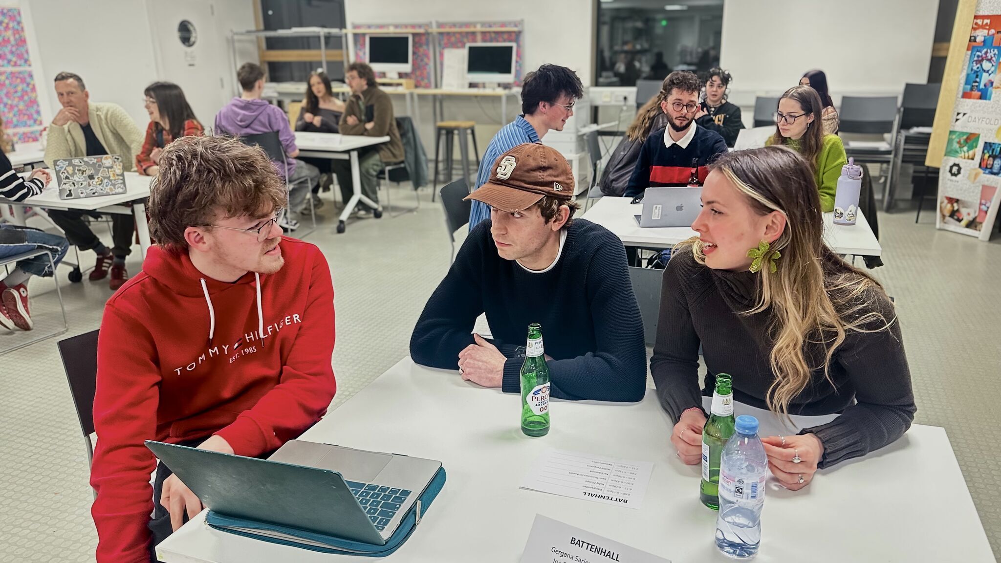 BA (Hons) Graphic Design students sitting at tables in seminar room, three in foreground - one in red hoodie with laptop, two others with water bottles and papers.