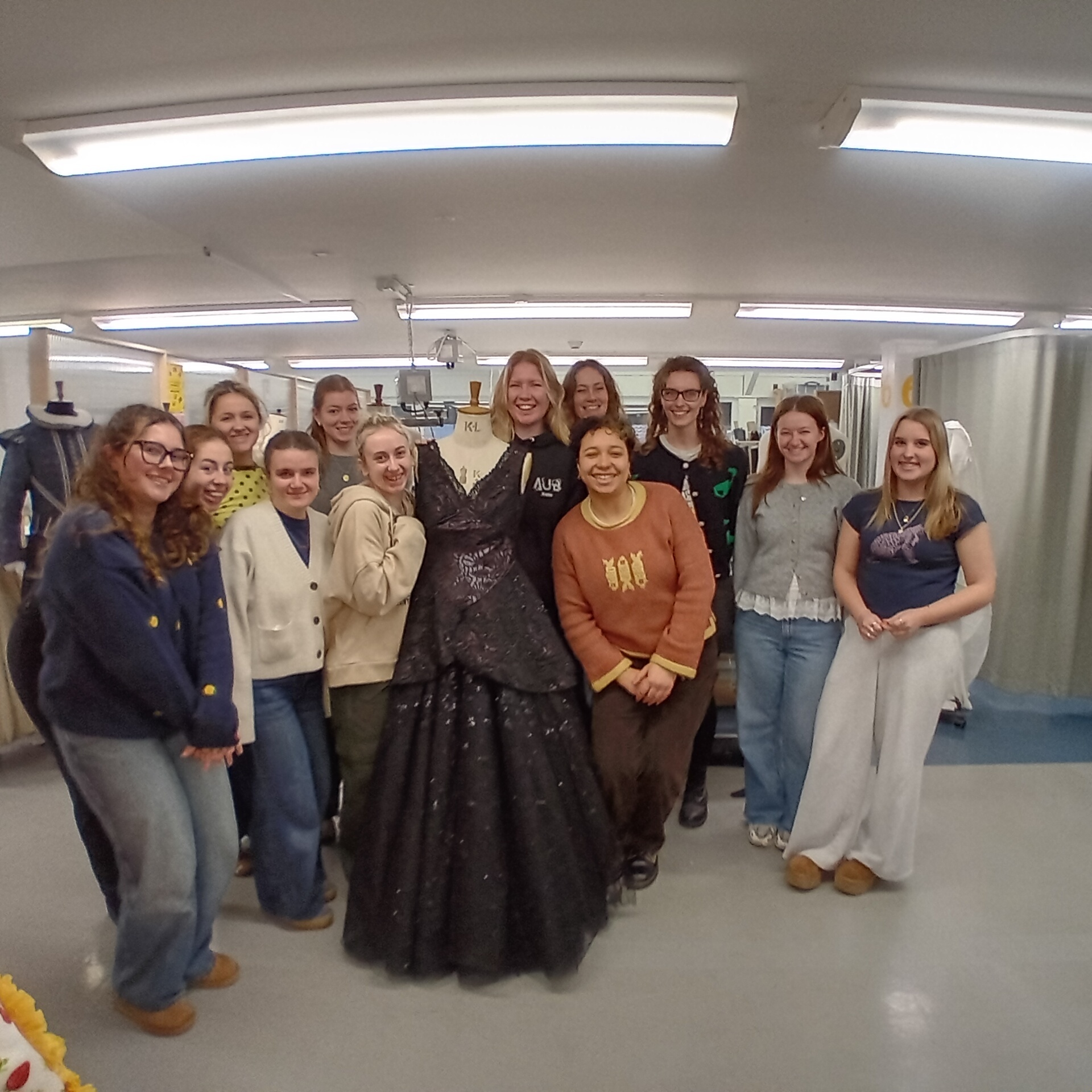 Group of BA (Hons) Costume students posing together in fluorescent-lit room with white walls and grey floor, wearing casual clothing and jumpers.