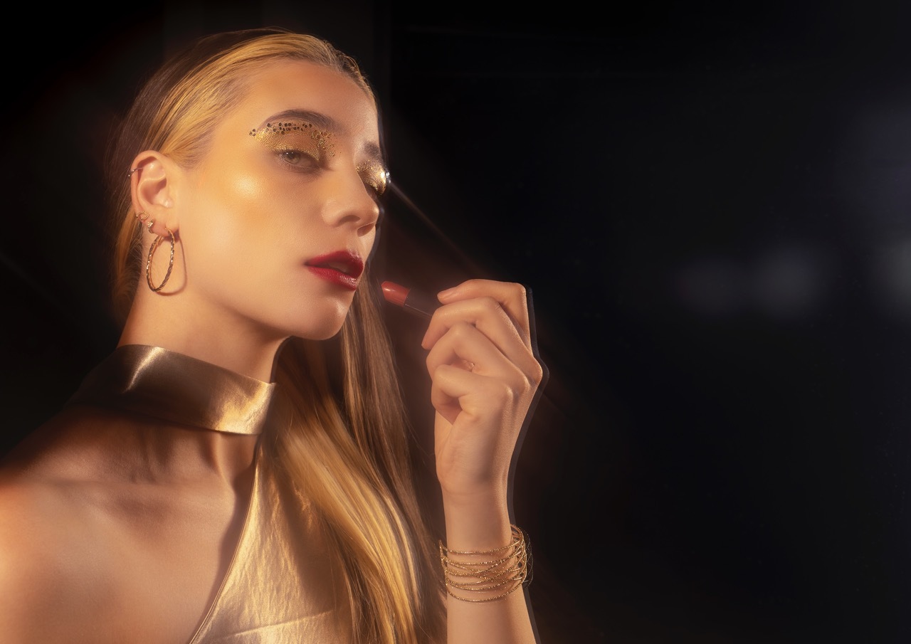 Blonde woman with gold choker necklace and hoop earrings, hand raised to lips applying red lipstick, warm lighting against dark background.
