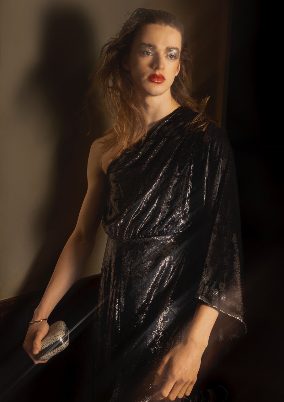 Woman with wavy blonde hair wearing black one-shoulder sequinned dress, red lipstick, holding small clutch bag against dark background.