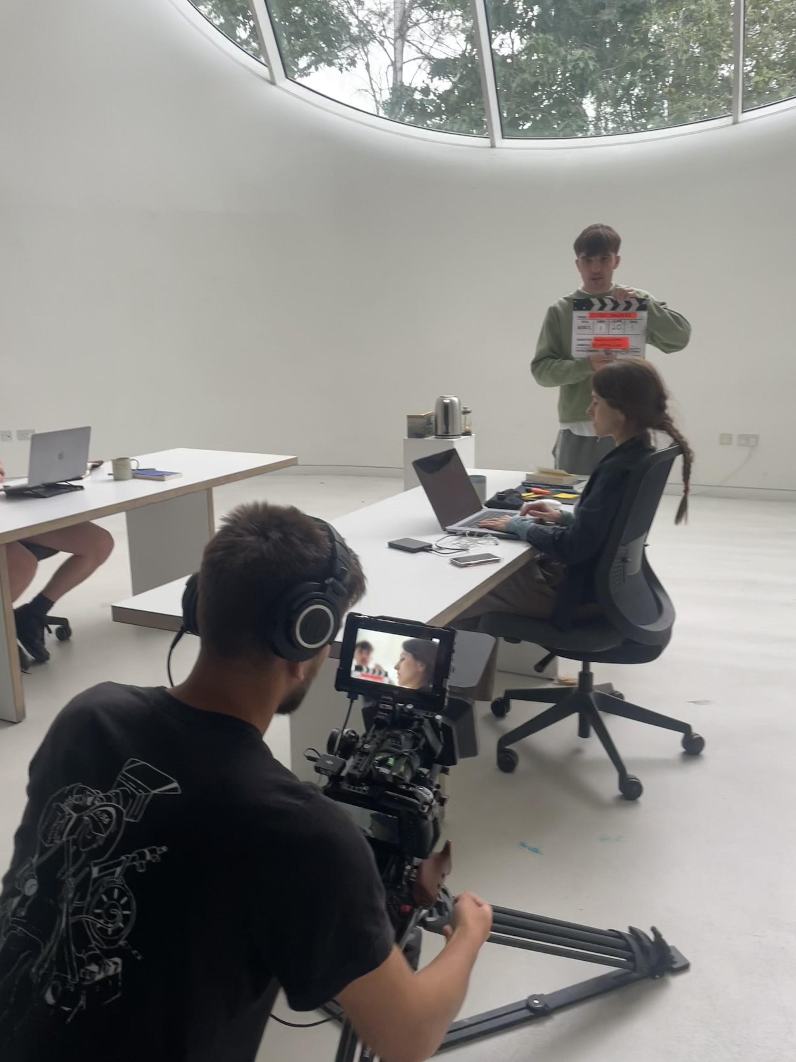 Film crew shooting in modern white office with curved ceiling windows. Cameraman operates equipment whilst woman sits at desk, man holds clapperboard.