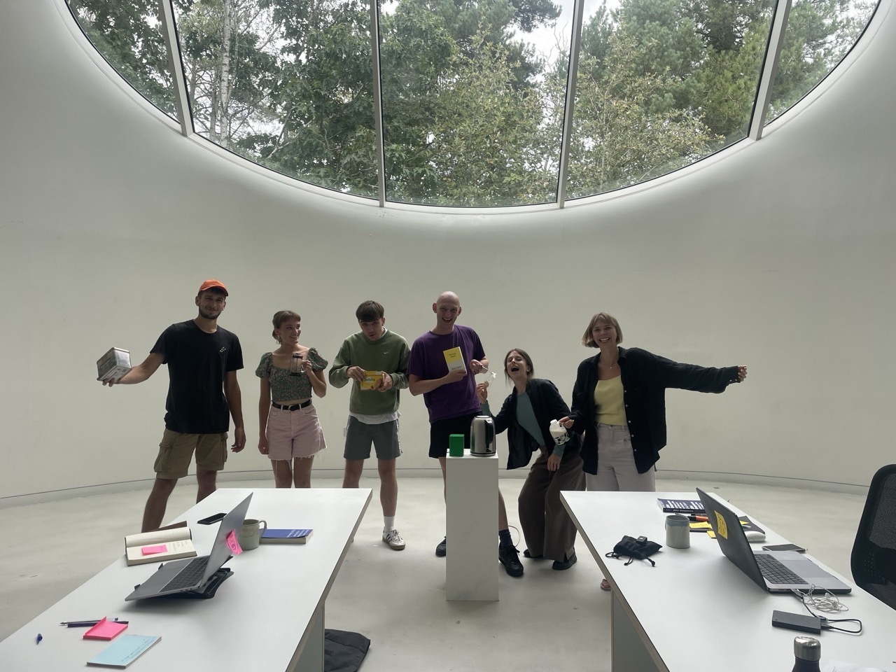 Group of six people standing with arms outstretched in white curved room with circular skylight showing trees above.