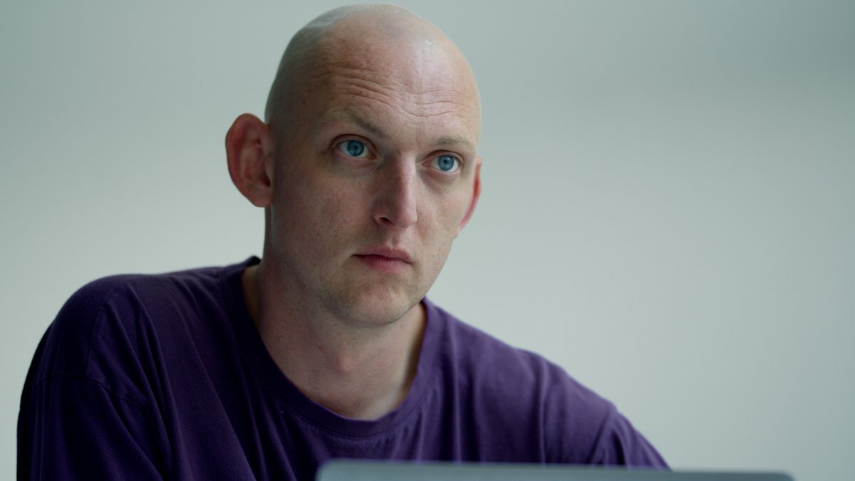 Bald man with light eyes wearing purple t-shirt against pale grey background, looking directly at camera.