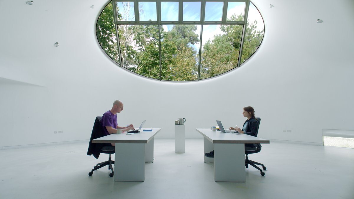 Modern office with two people at white desks beneath large oval skylight showing trees, minimalist white interior with polished floors.