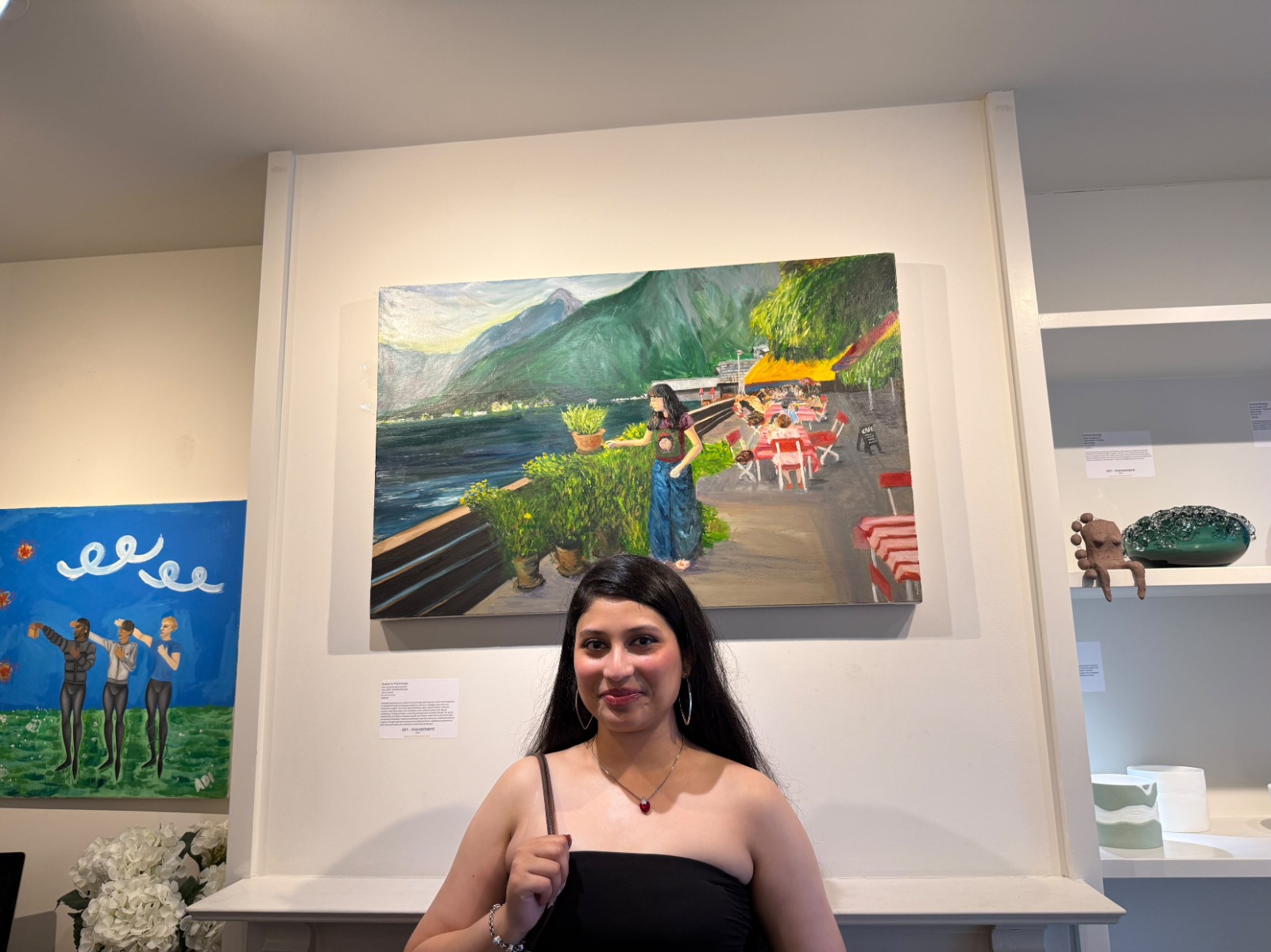 BA (Hons) Fine Art graduate S Mukherjee stands in front of a colourful painting of a landscape, featuring mountains and a waterfront scene with tables and chairs. The background shows a white wall with various art pieces displayed.