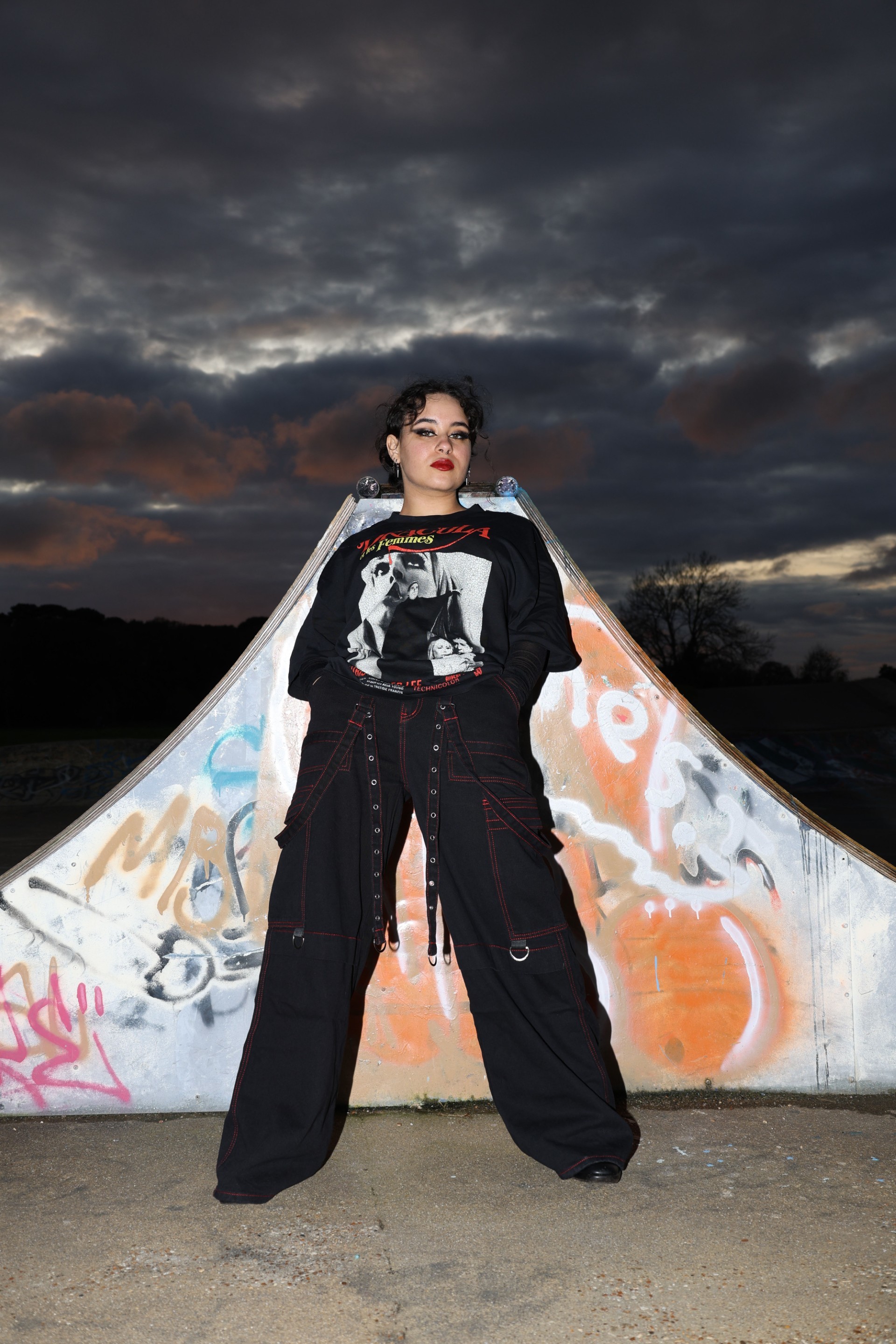 Kasandra Georgi posed in front of graffiti skate wall in black flashy clothing, dusk sky in the background.