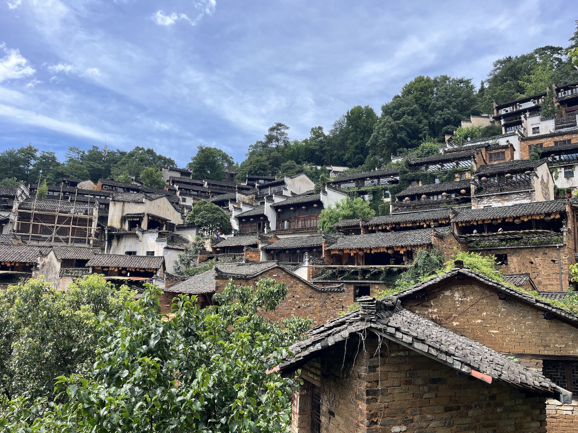 A hillside village of traditional stone houses with tiled roofs, set among dense green trees beneath a partly cloudy blue sky.