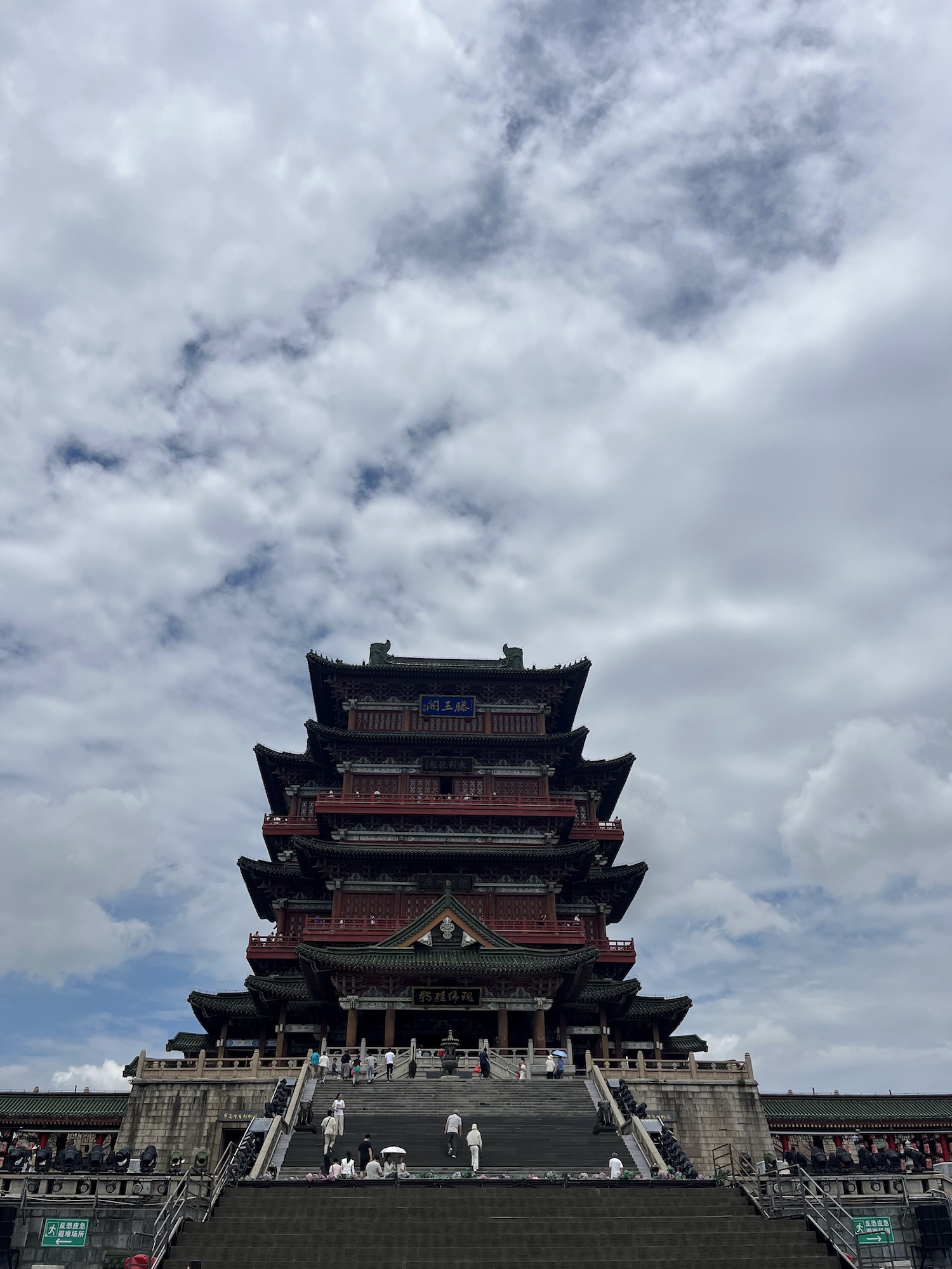 A multi-tiered traditional Chinese temple or pagoda with red and dark roofs stands at the top of broad stone steps under a cloudy sky.