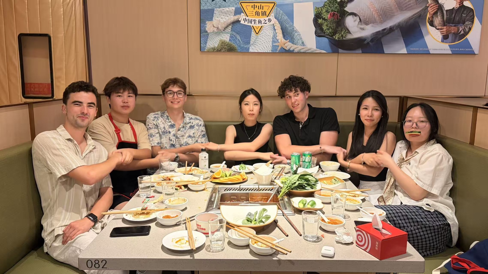 Seven people sit around a restaurant table with plates of food and drinks, in a booth beneath wall posters.