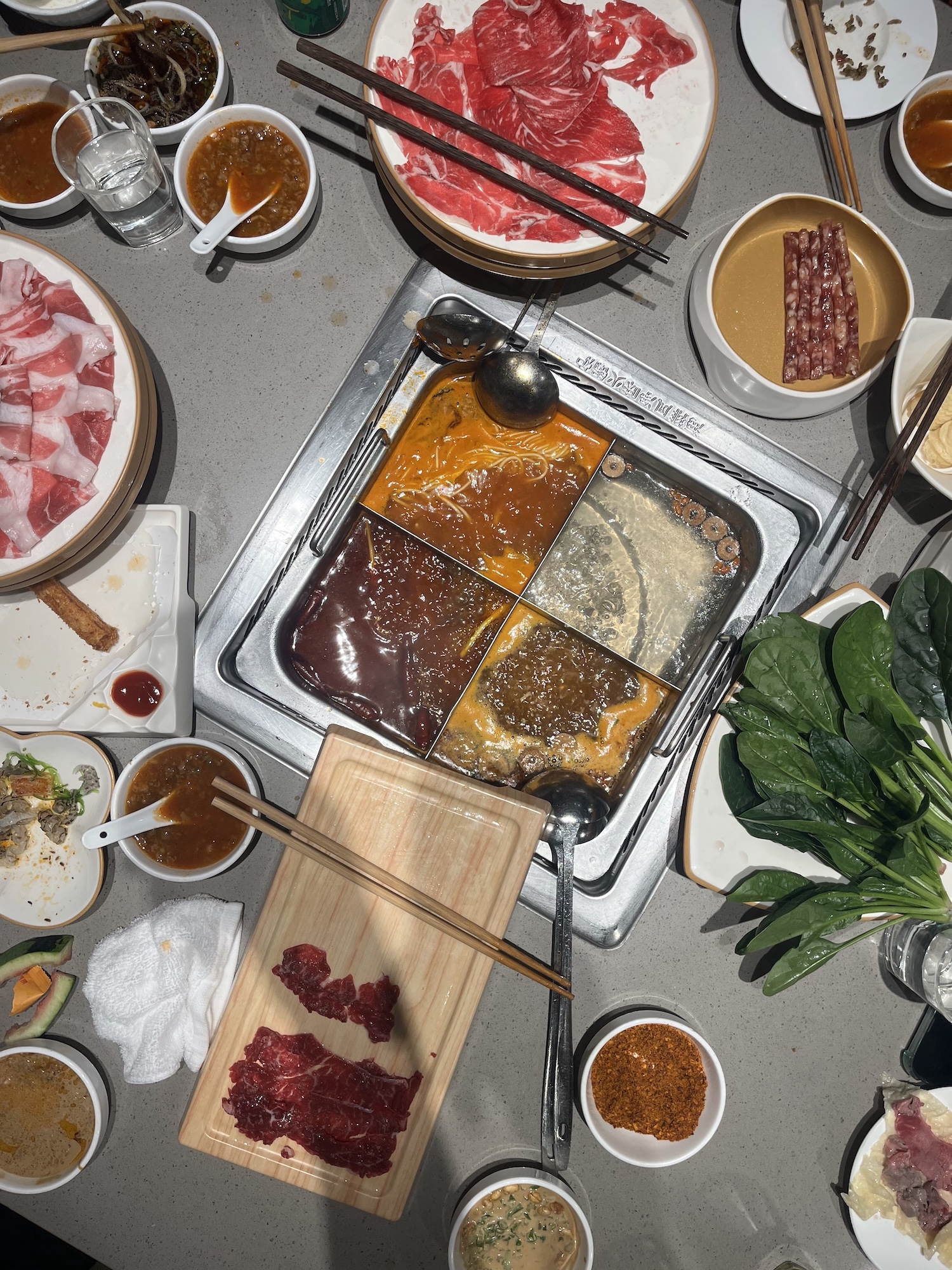 A hot pot meal with four divided broths in a table-top cooker, surrounded by plates of sliced meat, leafy greens, sauces and chopsticks.