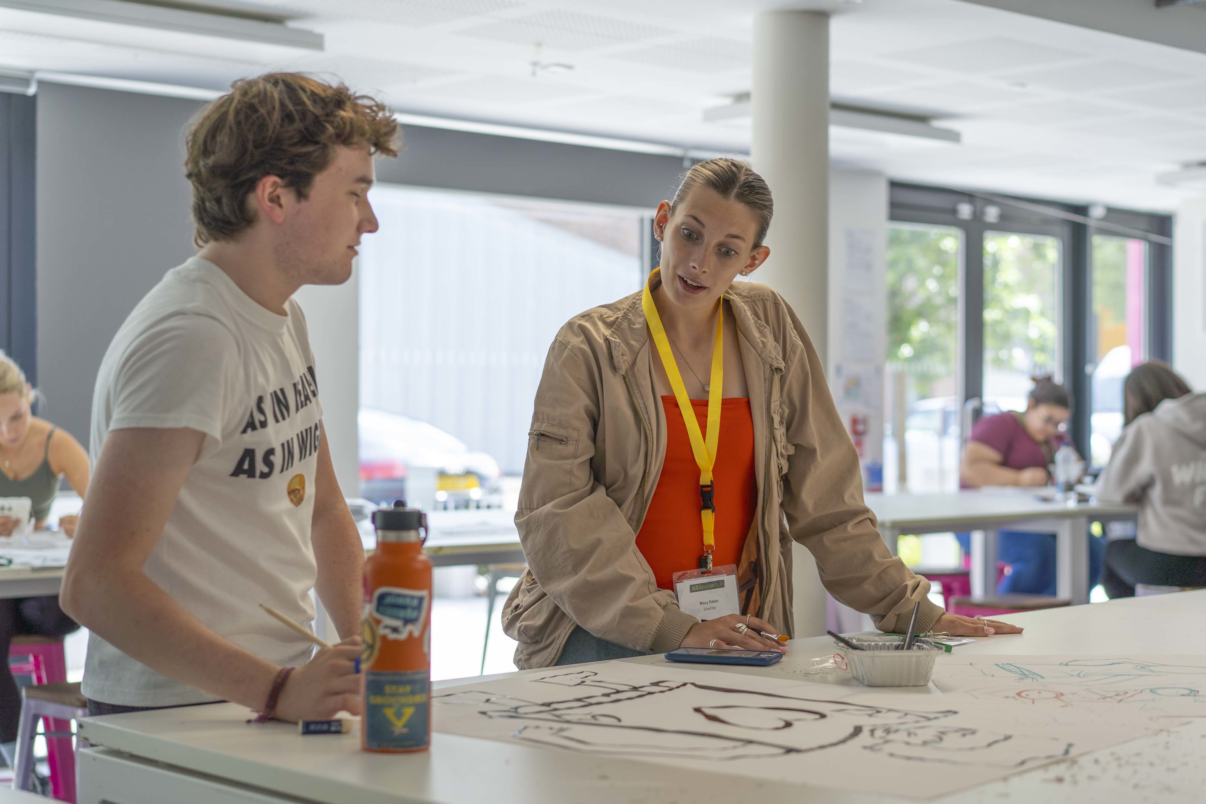 An AUB Outreach Ambassador standing with a young person at a table in an AUB studio discussing an artwork.