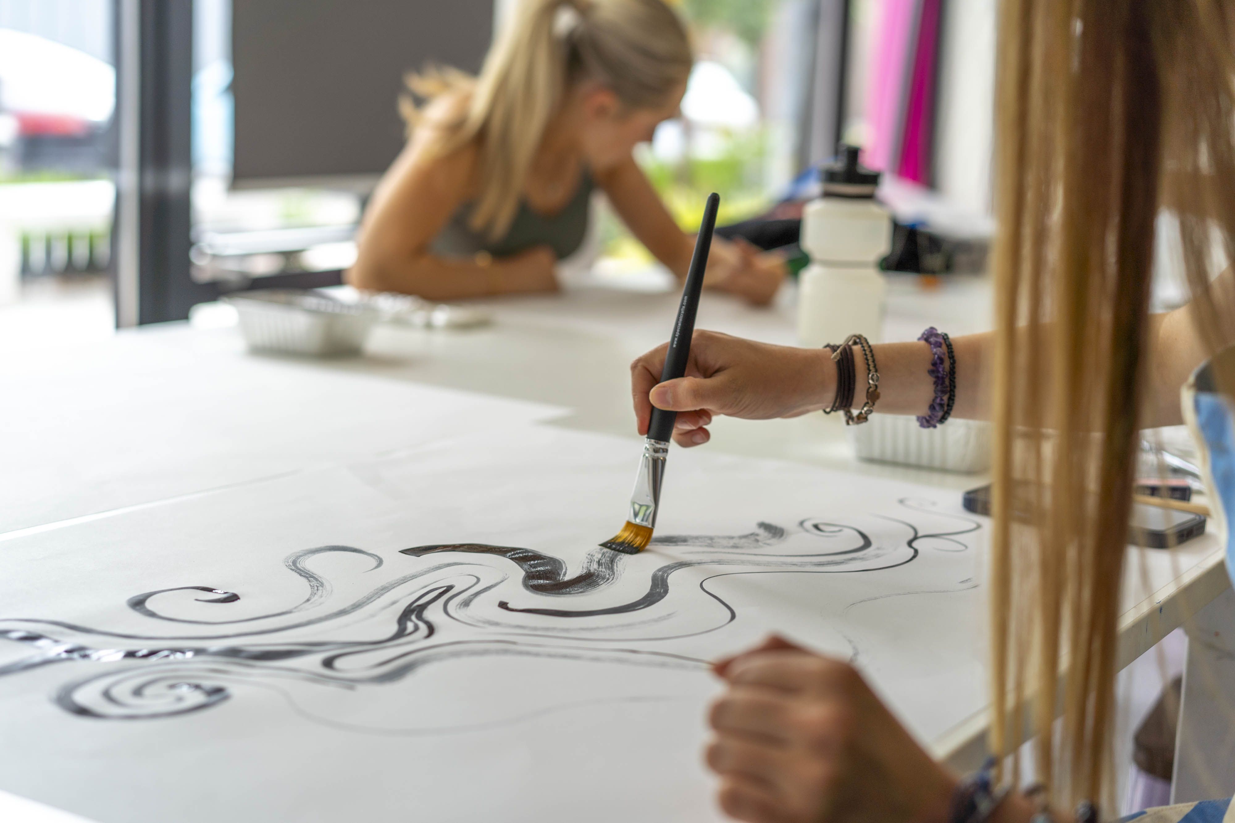 A close-up of a hand holding a paintbrush, applying black paint to a white canvas. In the background, a woman is working at a table, partially visible, focused on her own artwork.