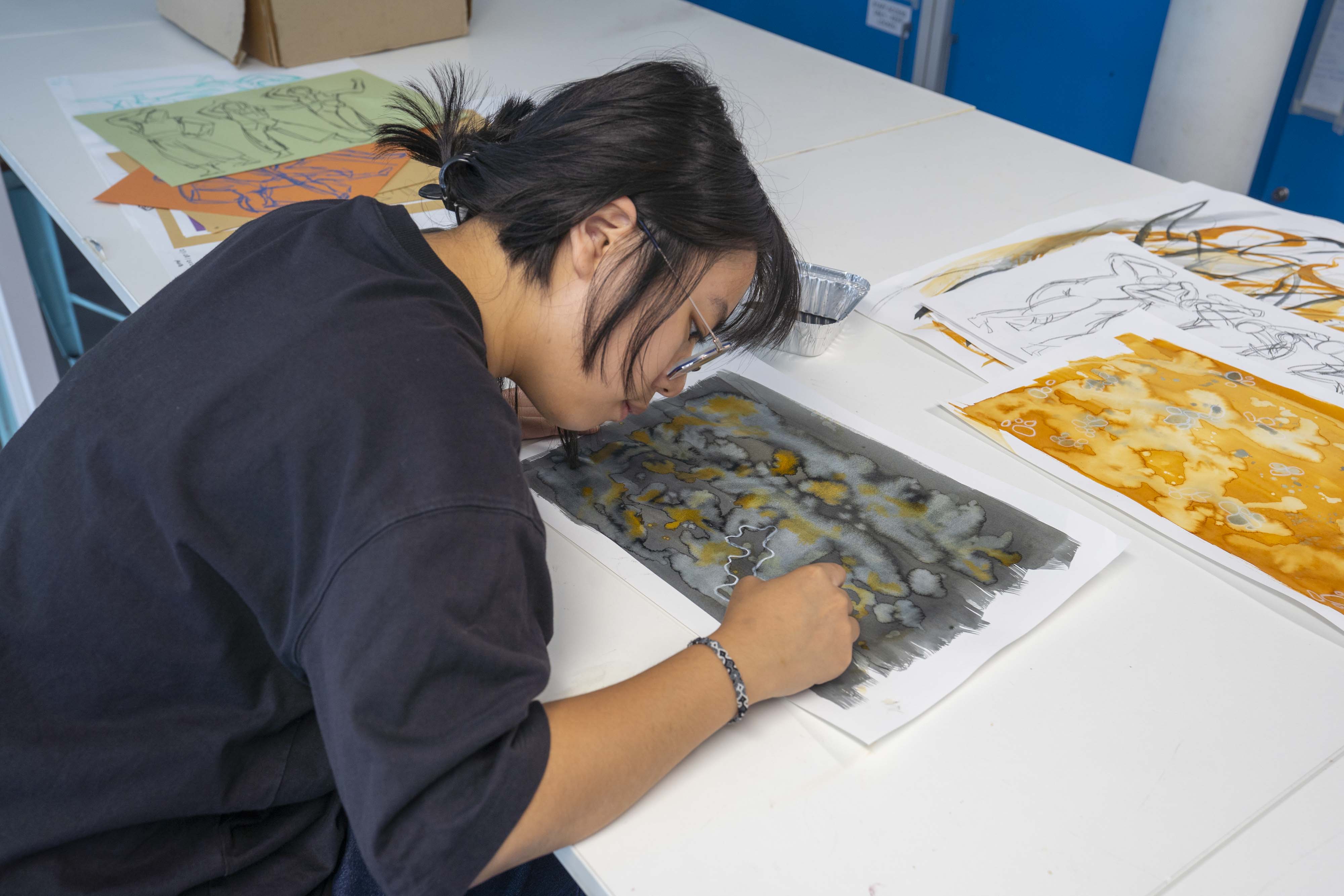 A young person with long dark hair is focused on painting at a table covered with various artwork. They are using a brush to add details to a painting featuring a grey background with yellow and orange elements, surrounded by other sheets of art.
