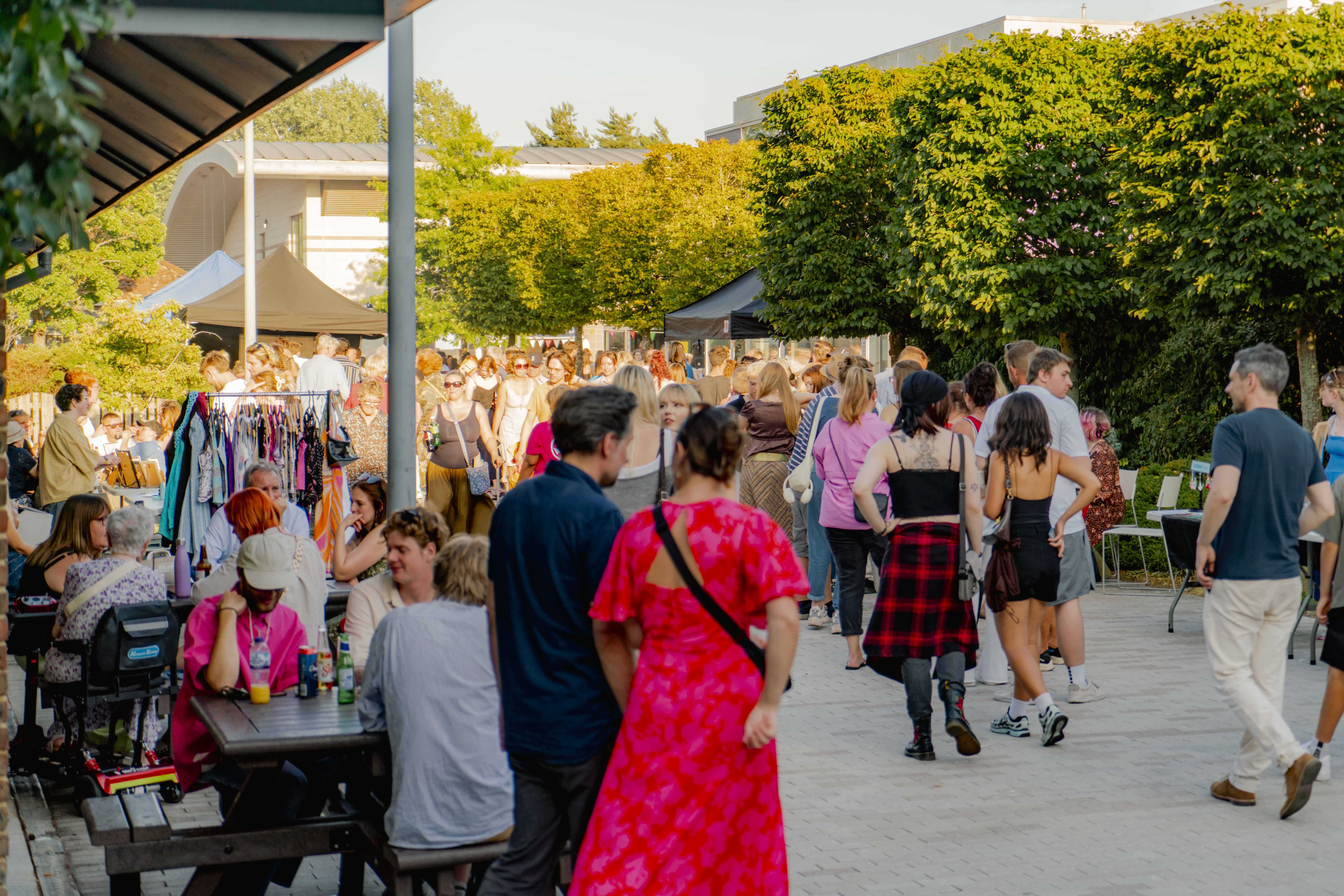Large crowd of people in AUB courtyard on a sunny evening, woman in bright pink-red dress blurred in front, trees above.