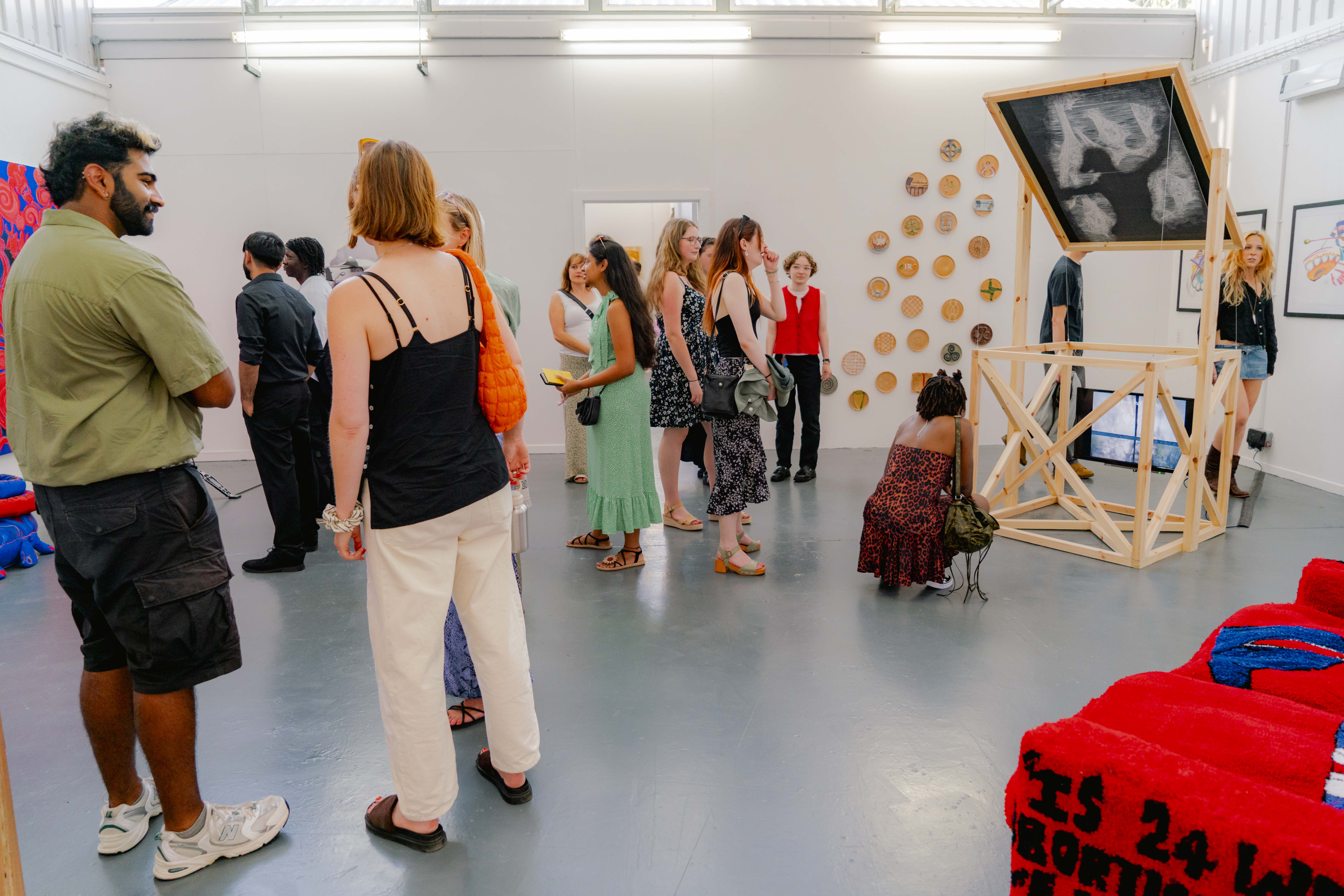 People stood talking and looking at in a white studio space, wood frame structure and red fabric car to the right.