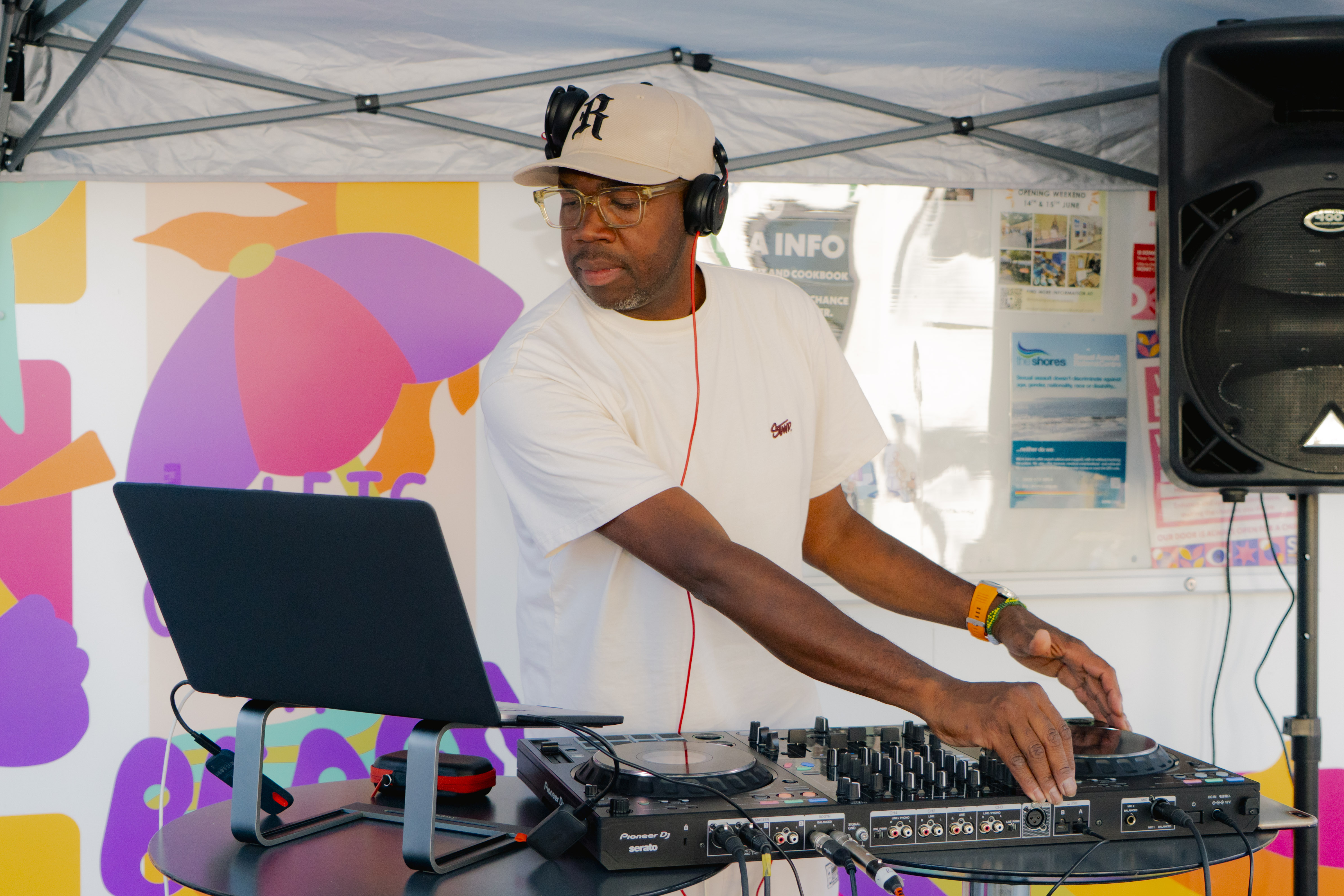 DJ Will on the DJ wearing a cap and headphones, looking at a laptop and interacting with equipment, colourful background.