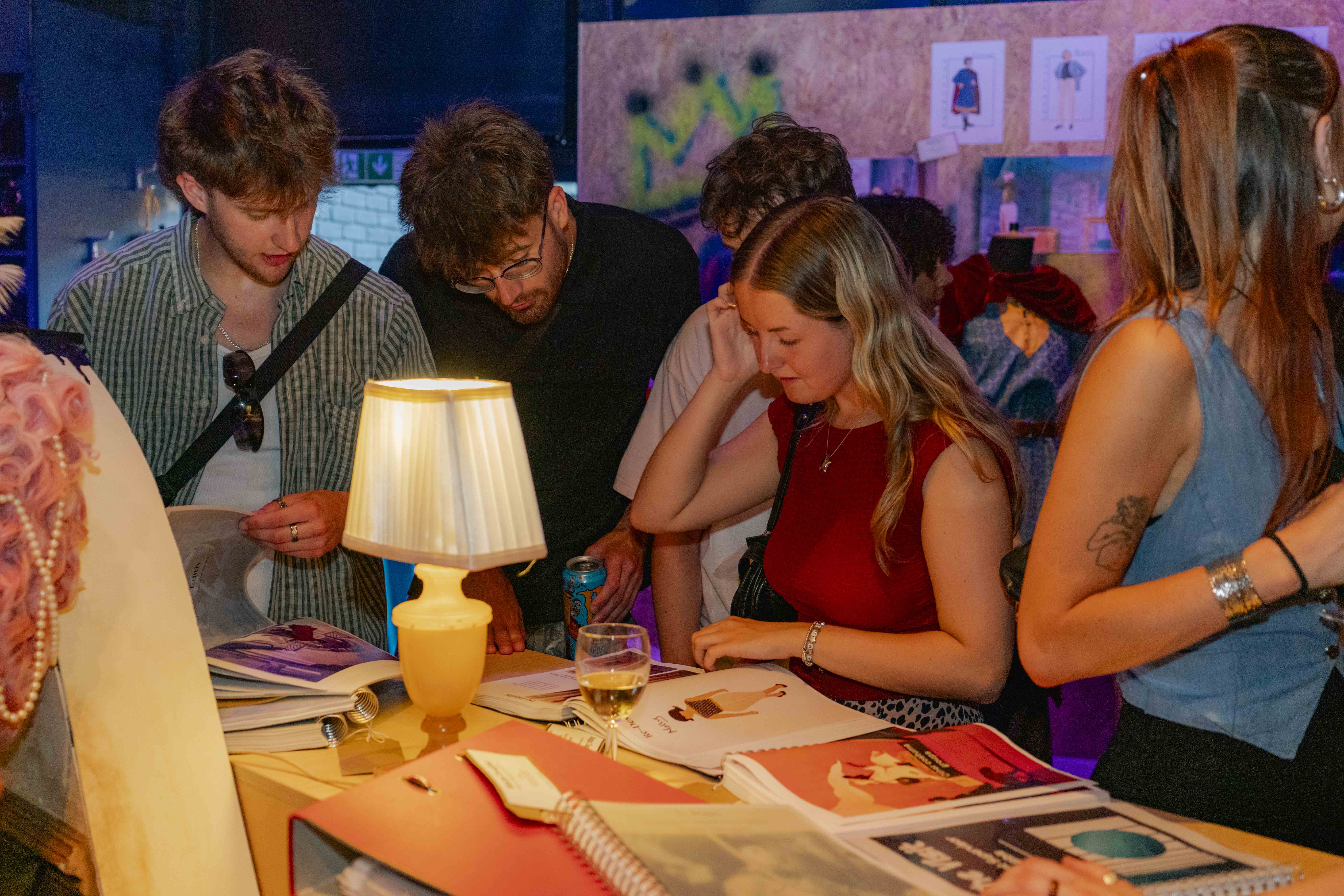 People looking at work on a red table, illuminated by a warm table lamp, dark purple background with wood display wall.