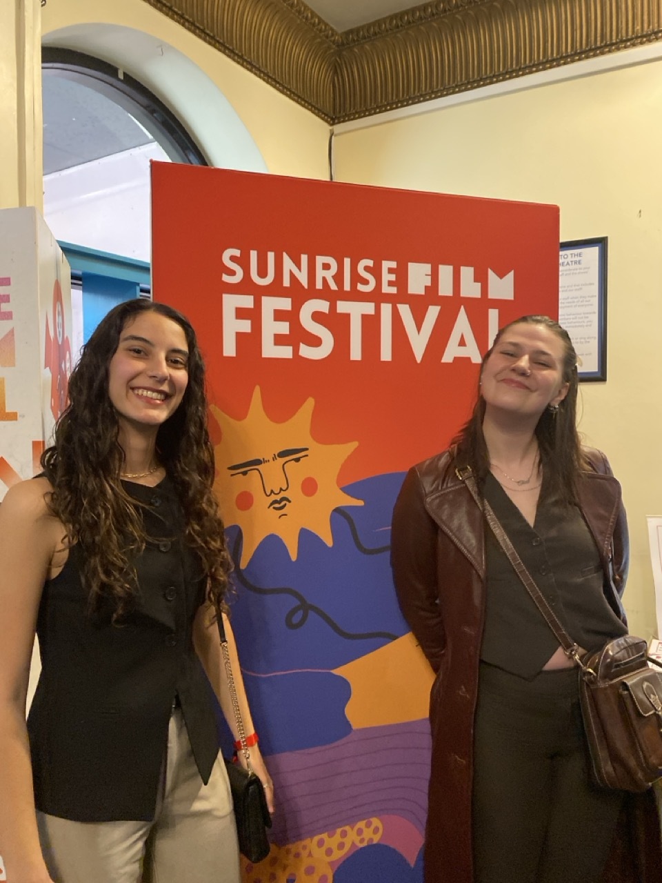 Two women standing beside red Sunrise Film Festival banner with colourful sun design in yellow, orange, blue and purple.