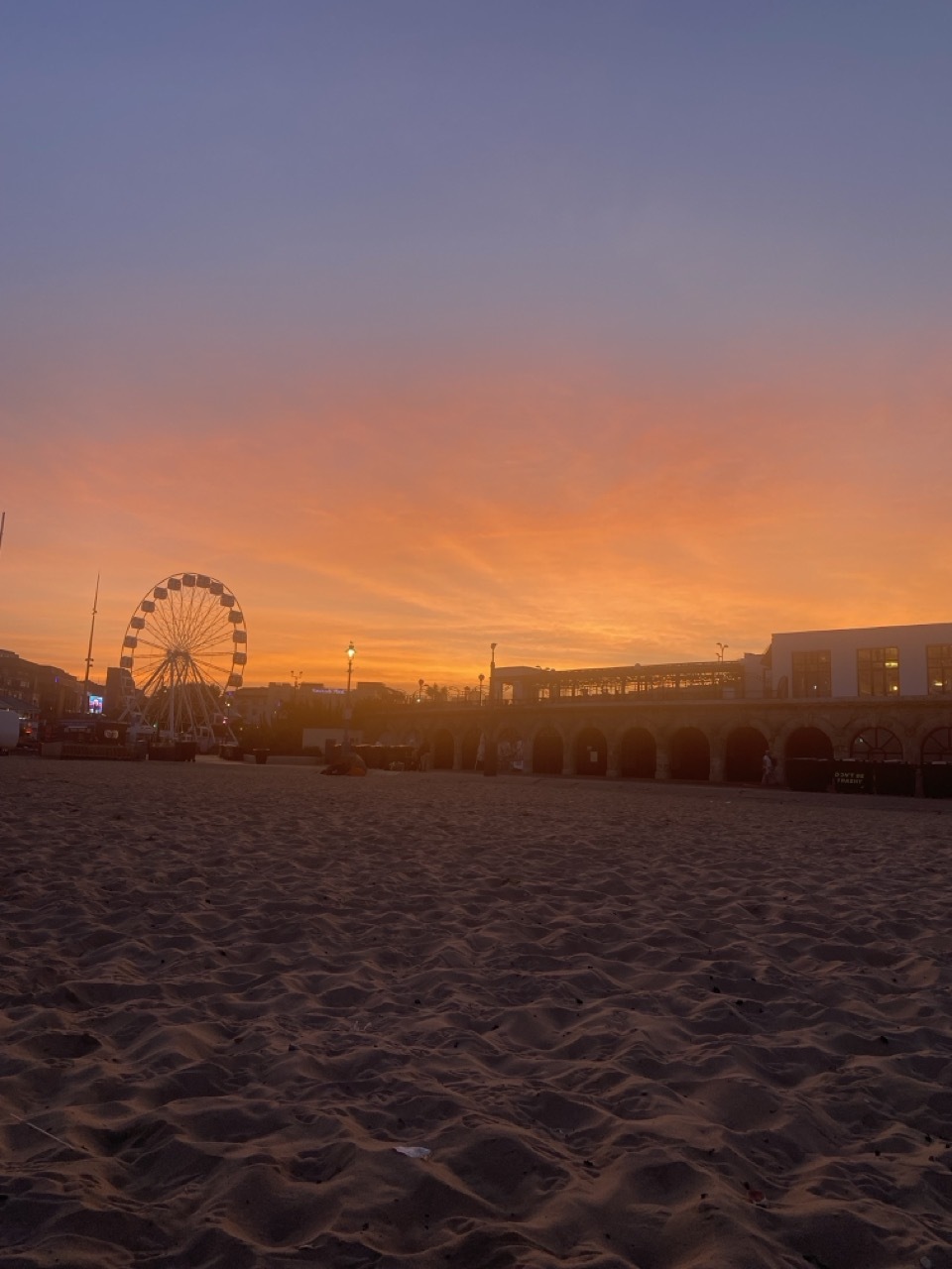 Sandy beach in foreground with Ferris wheel and buildings silhouetted against orange and purple sunset sky.