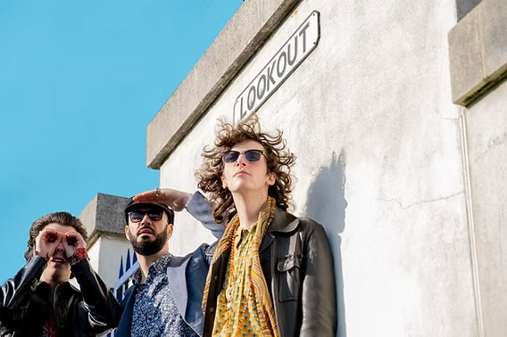 Three people wearing sunglasses, one with his hands cupped over his eyes like binoculars, the other shielding his face with one hand, looking off into the distance, standing under a street sign that says "Lookout".